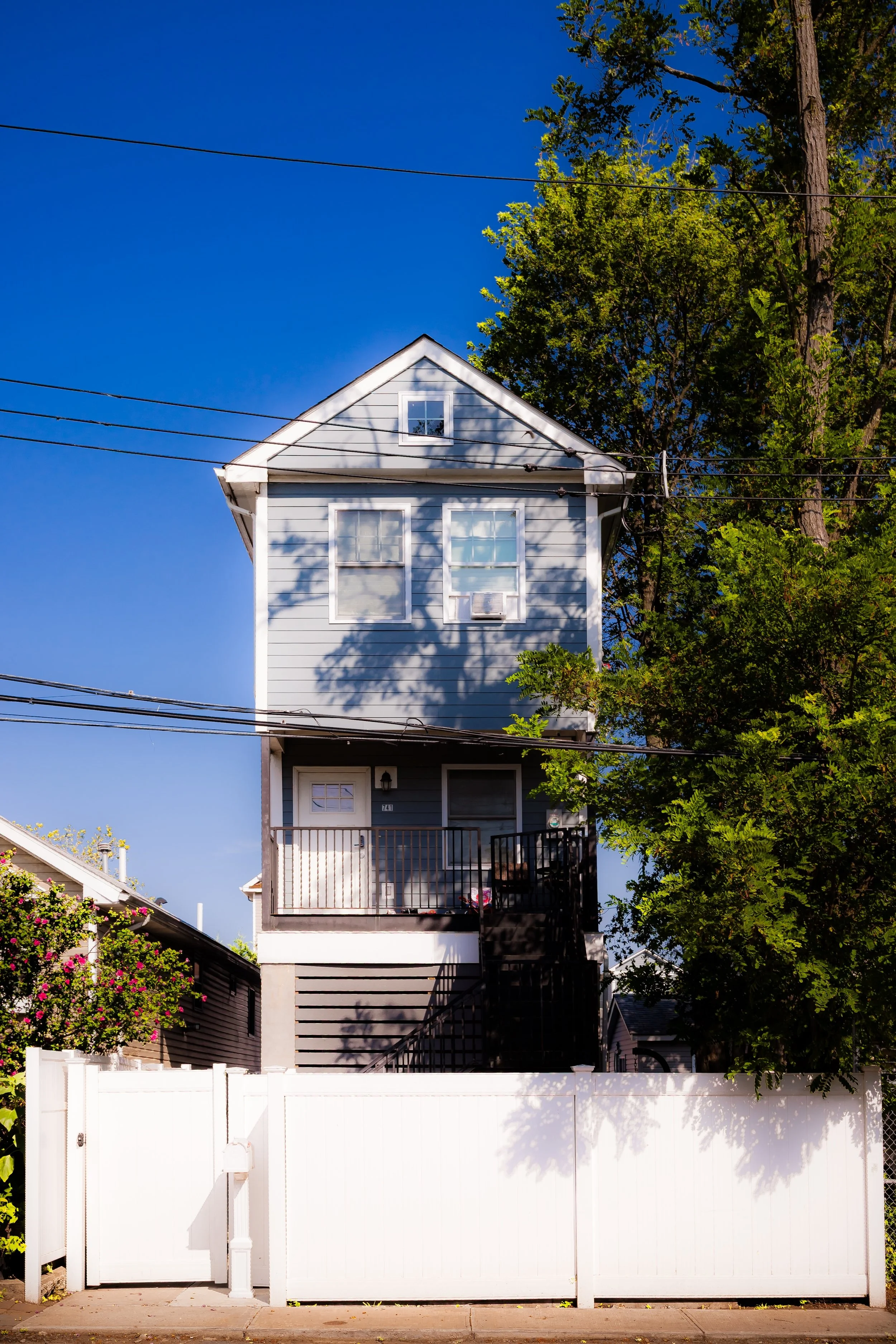 Three-story blue house with white trim, a small front porch, and a white fence. There are trees and clear blue sky in the background.