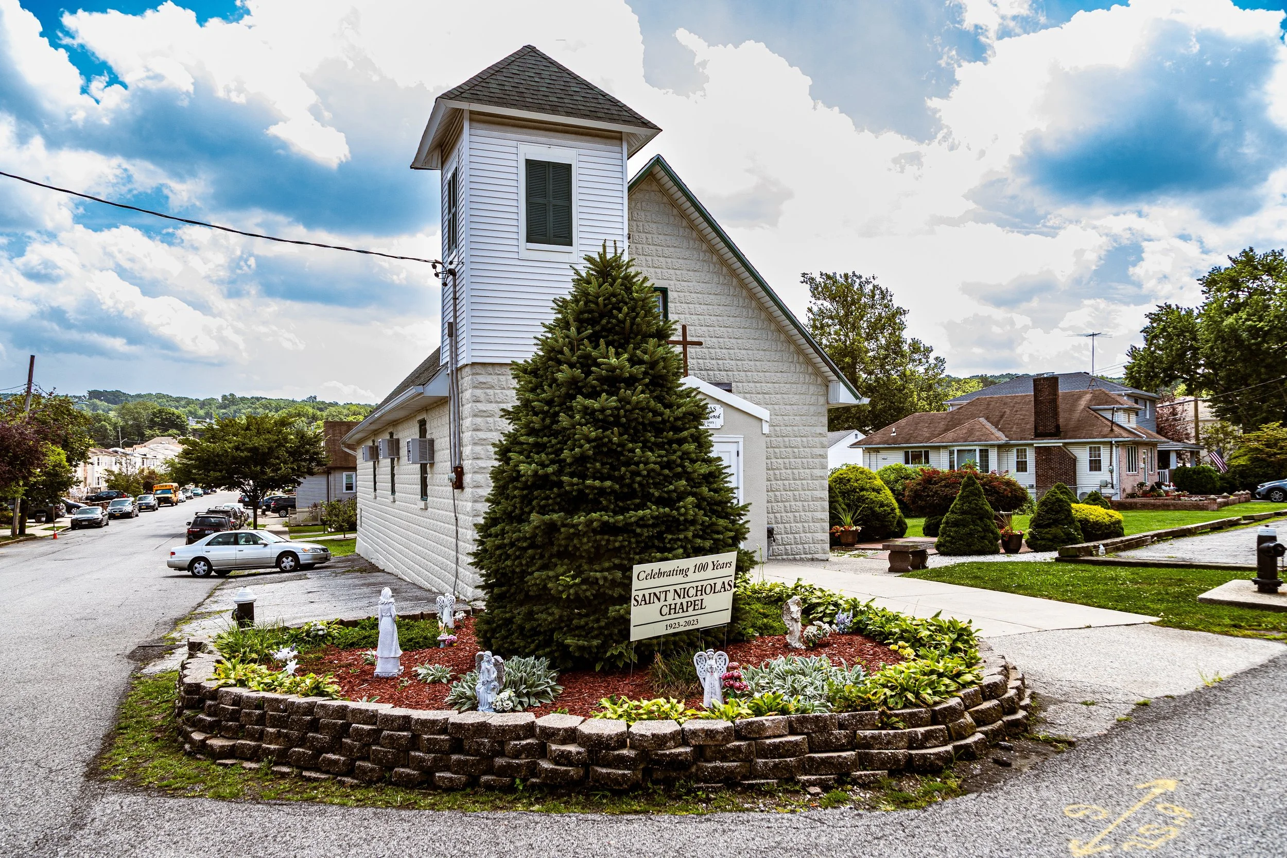 A church with a tall steeple, surrounded by a cul-de-sac and a garden with a sign celebrating 100 years of Saint Nicholas Chapel from 1923 to 2023, with a tree and angel statues in front, under a sky with scattered clouds.
