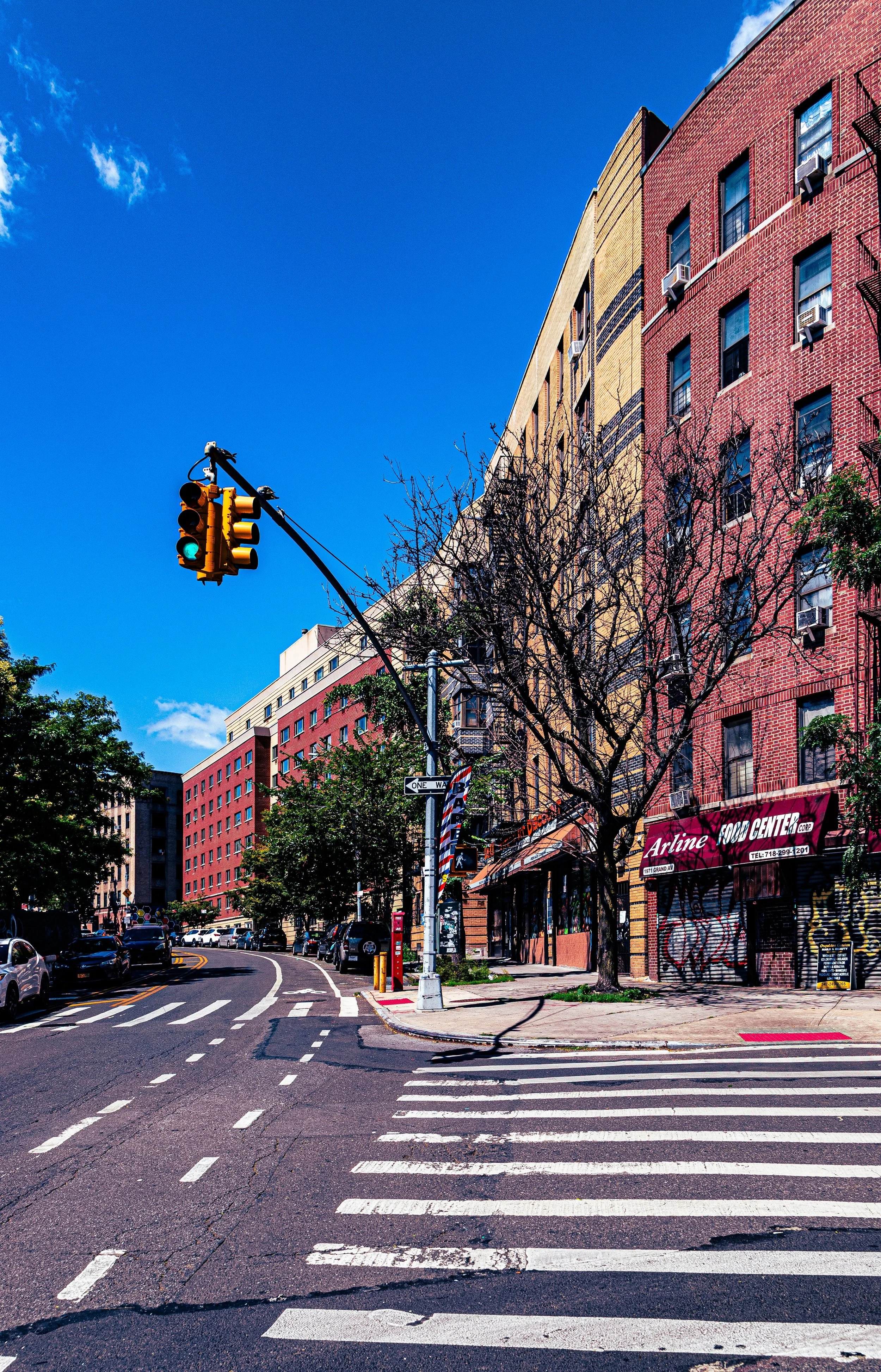City street scene with colorful multi-story buildings, cars parked along the street, a traffic light blinking green, leafless trees, a crosswalk, and a blue sky with a few clouds.