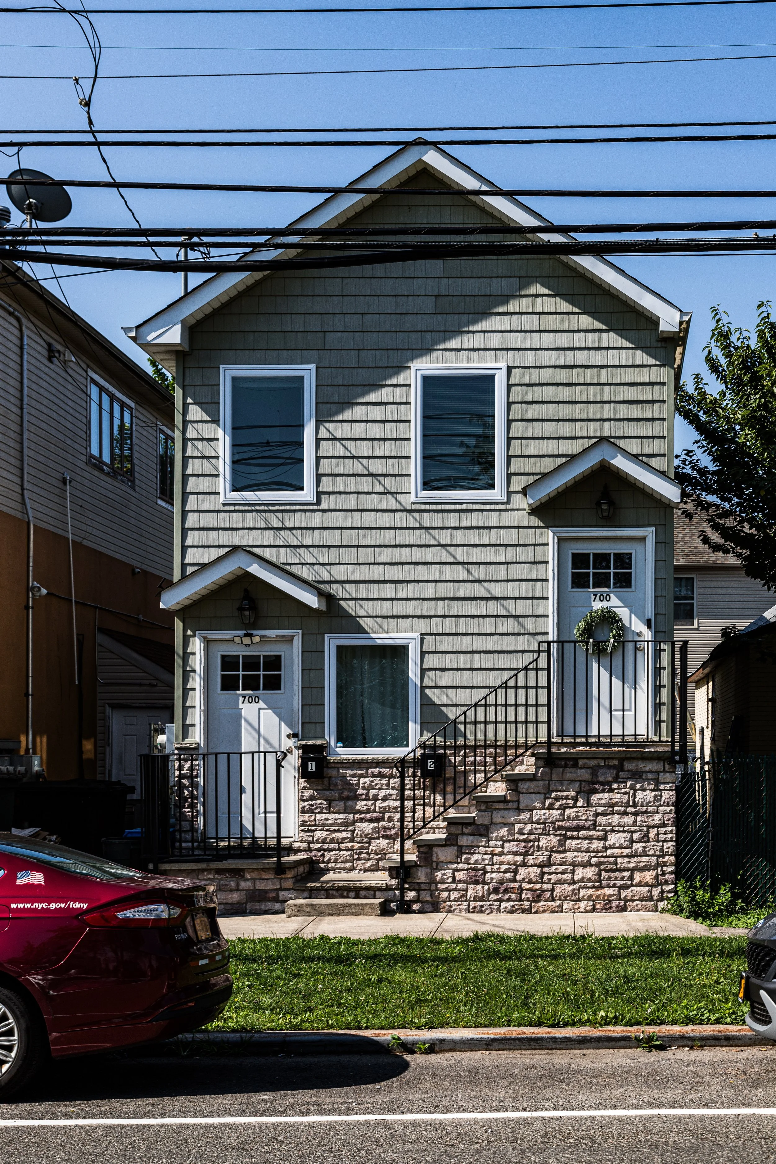 A two-story house with green shingle siding, two front doors with small porches and wreath decorations, stone foundation, and multiple windows, located on a city street with parked cars and overhead power lines.