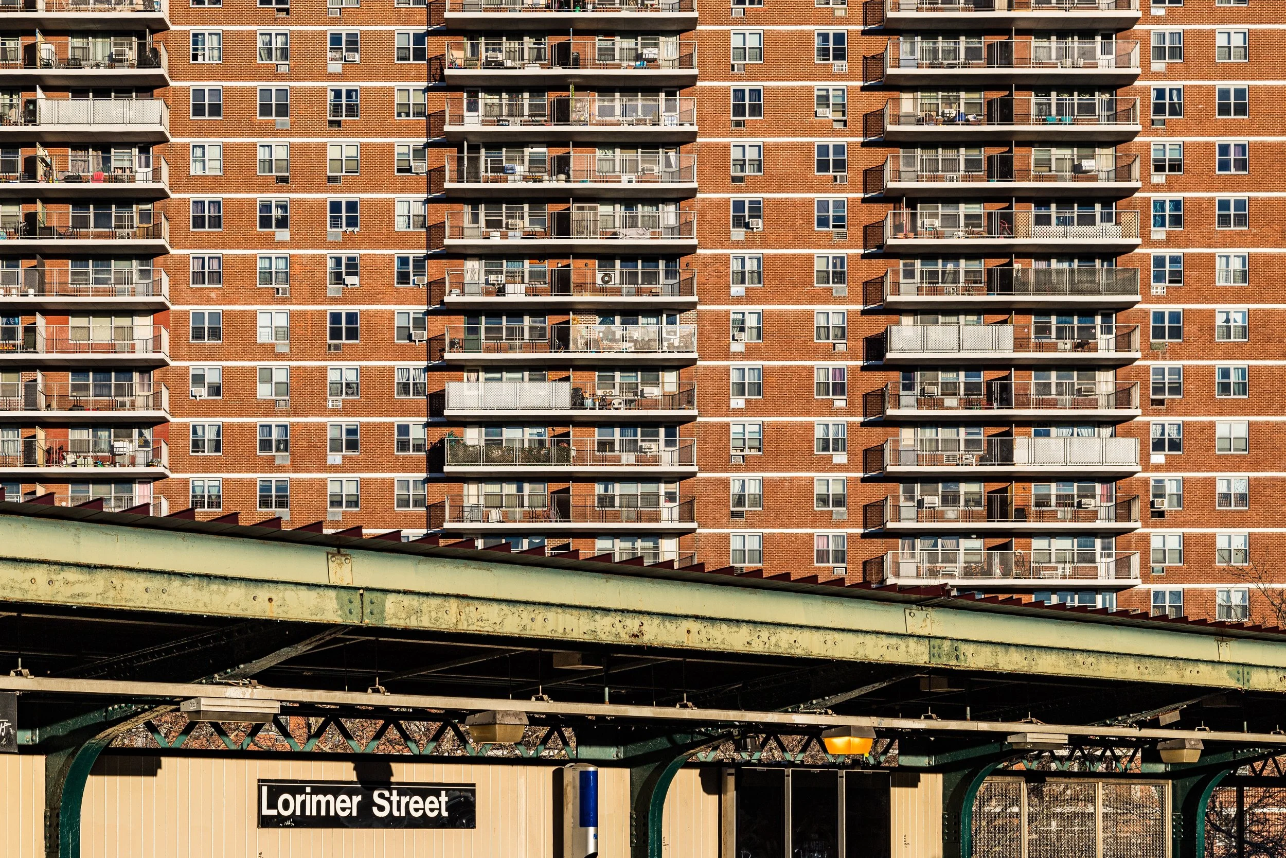 A tall red brick apartment building with multiple balconies on each floor, and a green metro station platform in the foreground with a Lorimer Street sign.