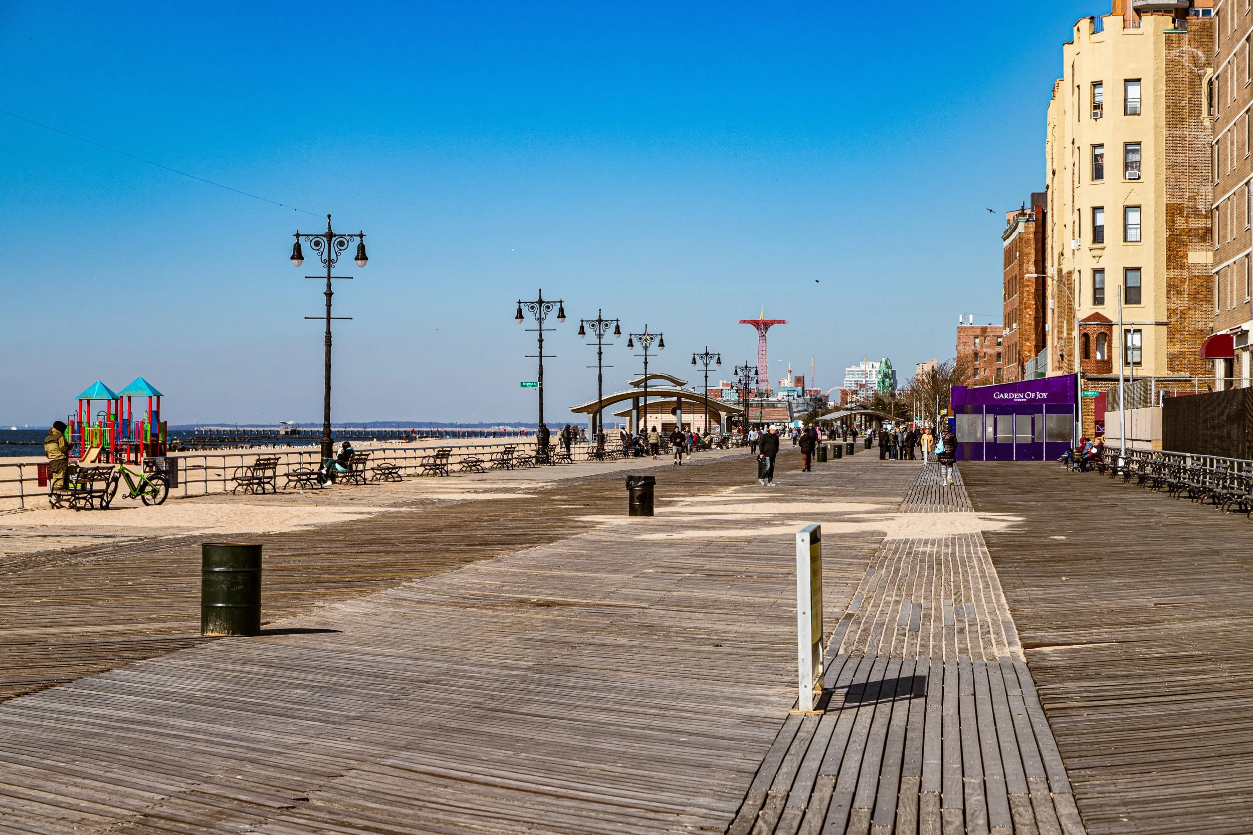 Empty boardwalk at a seaside pier with benches and street lamps, colorful playground equipment, and buildings in the background under a clear blue sky.