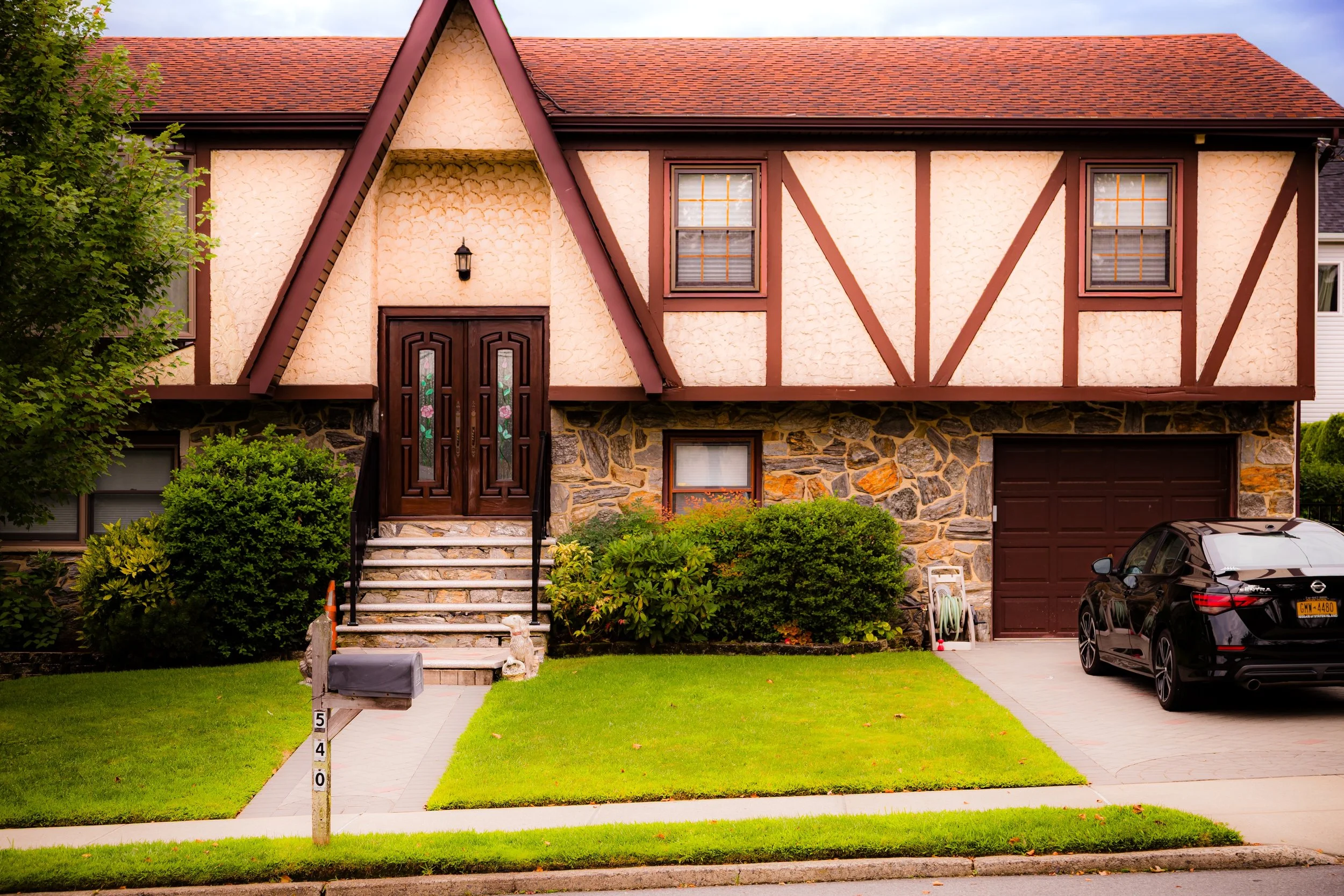 A two-story house with a Tudor-style facade, stone and stucco exterior, red tile roof, front steps, a black mailbox, and a driveway with a black car parked.