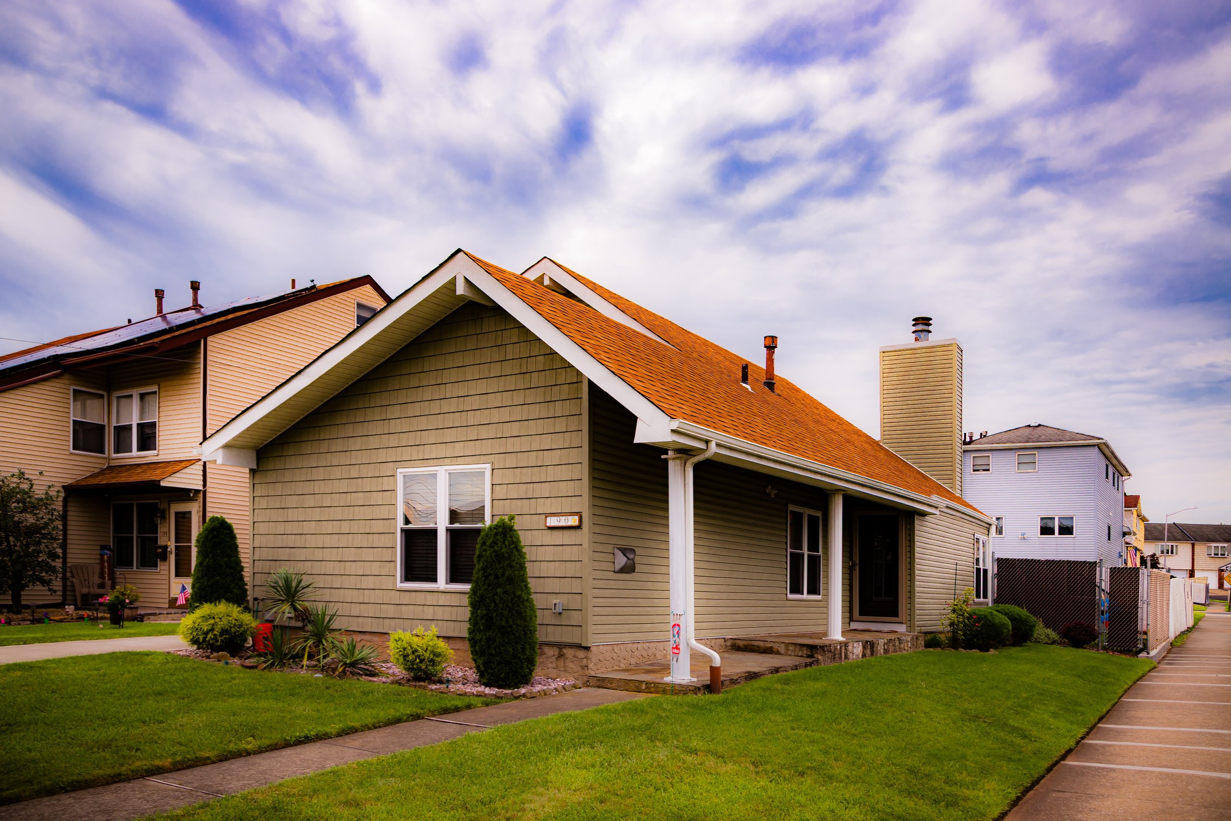 A house with beige siding and orange roof shingles, with a well-maintained lawn and landscaped bushes, under a partly cloudy sky.