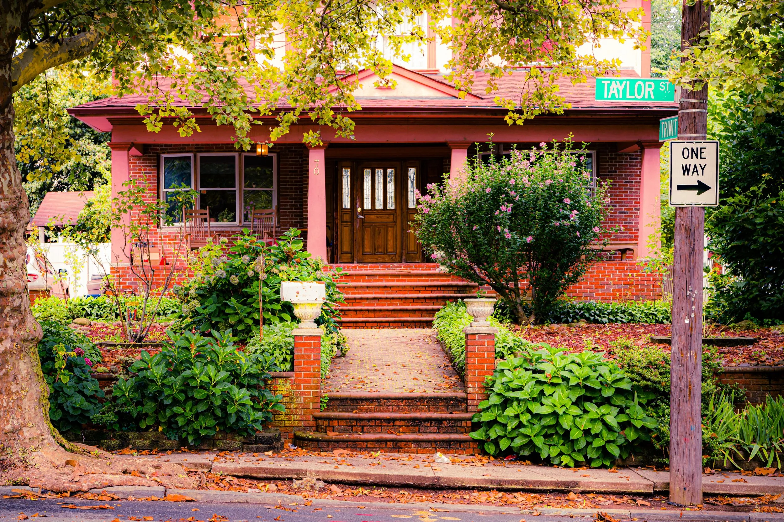 A front yard with lush green plants and bushes, brick steps leading to a wooden front door, and a house with brick walls and pink columns. There are street signs indicating the intersection of Taylor Street and Transit Street with a one-way sign pointing right.
