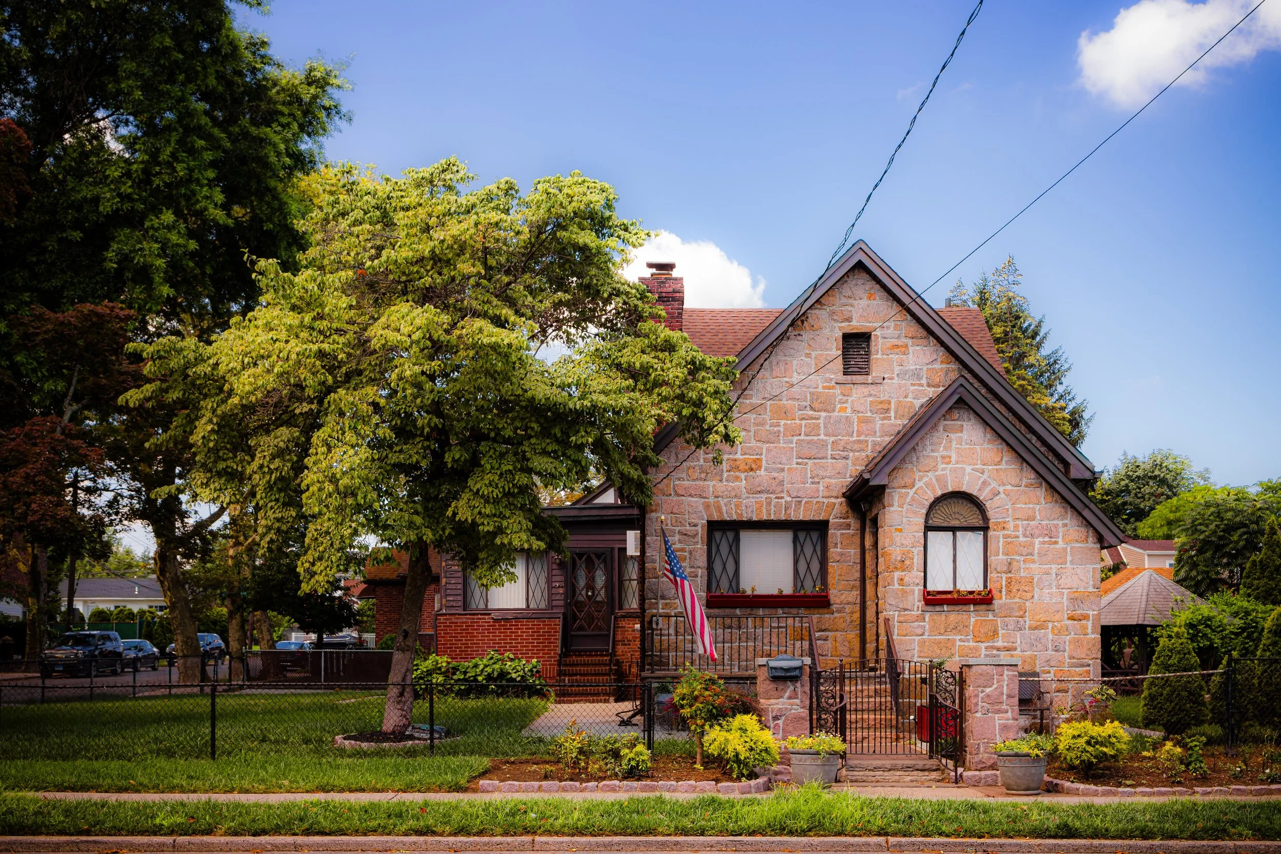 A brick house with a gabled roof, surrounded by trees and a small garden, with an American flag hanging near the front door.