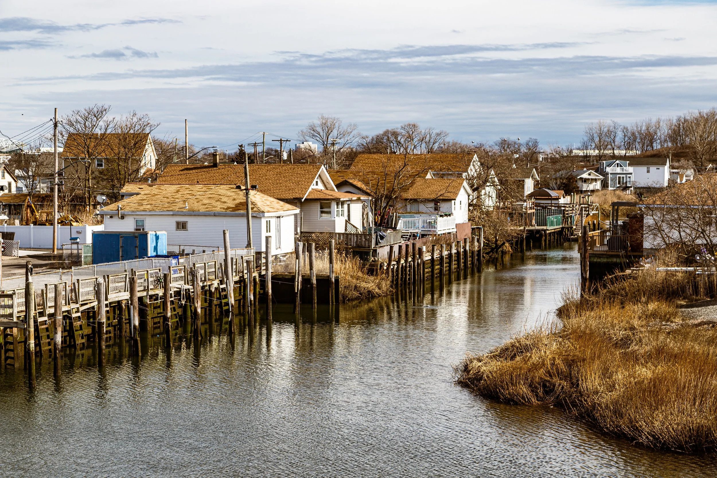 Residential houses along a waterway with wooden docks, some houses on stilts, and a cloudy sky.