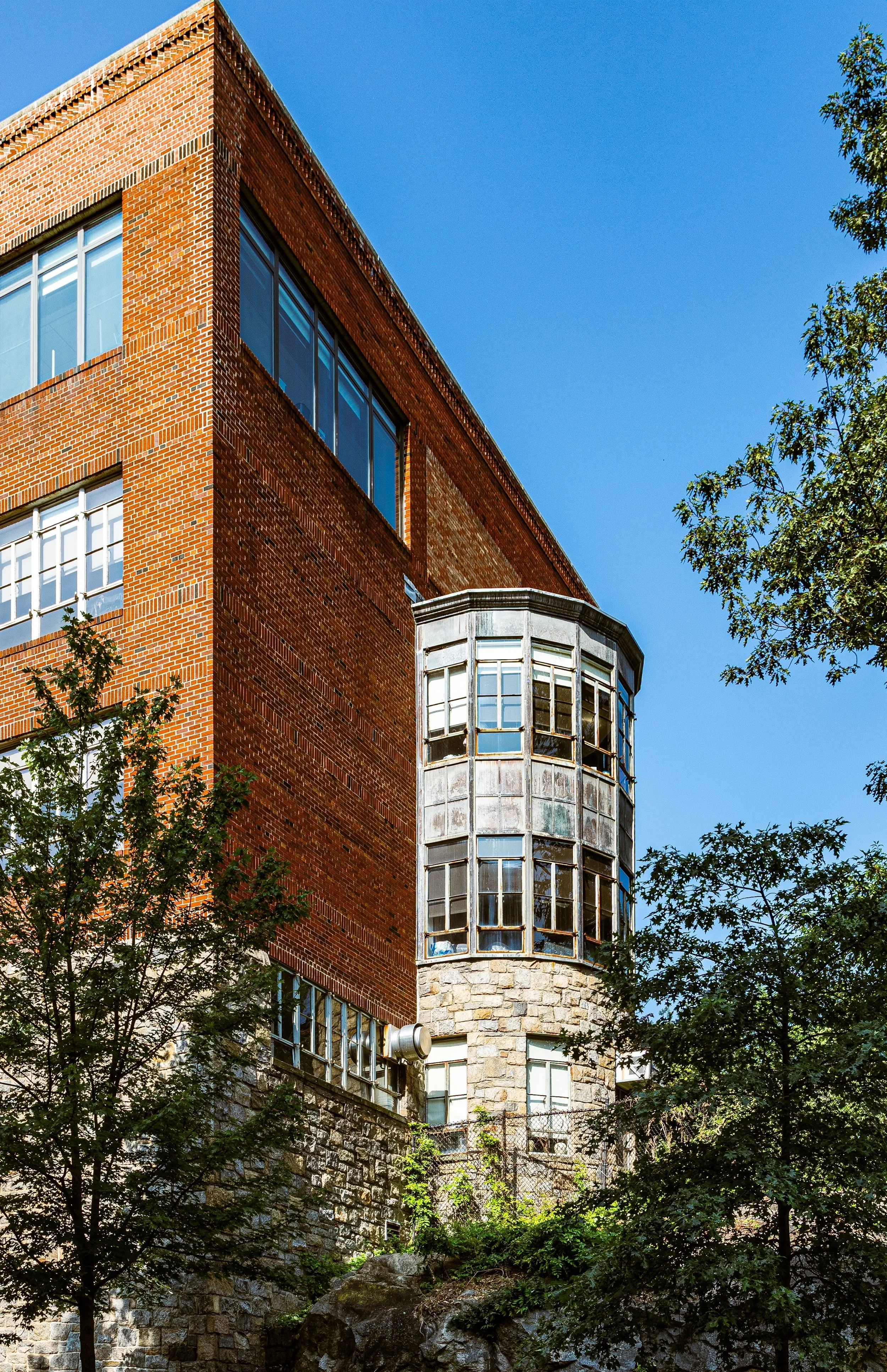 A close-up view of a multi-story brick building with large windows, featuring an enclosed stone turret with glass windows, surrounded by trees under a clear blue sky.