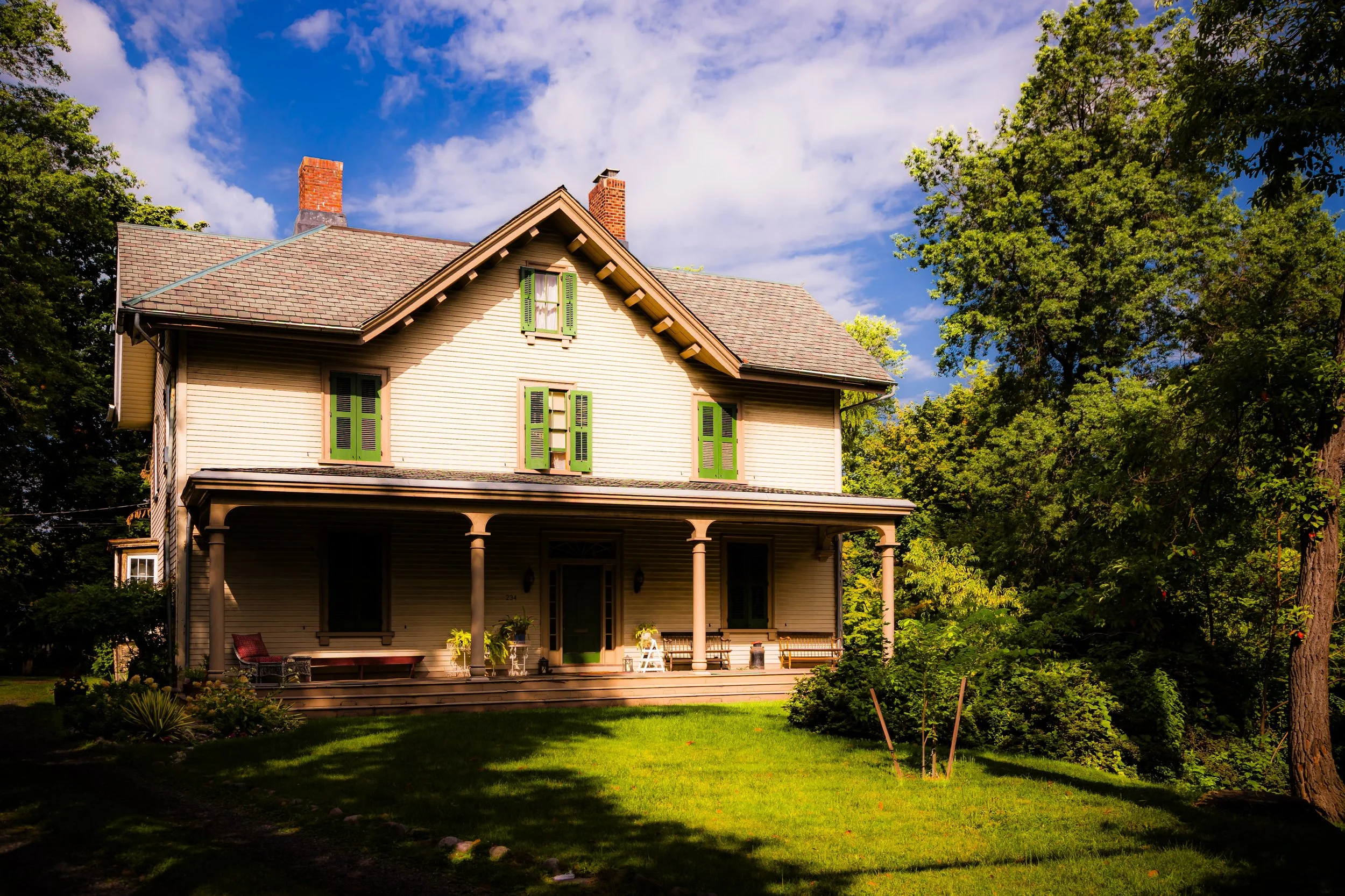 A large two-story house with cream-colored siding, green window shutters, a front porch with columns, and a well-maintained lawn surrounded by trees under a partly cloudy sky.