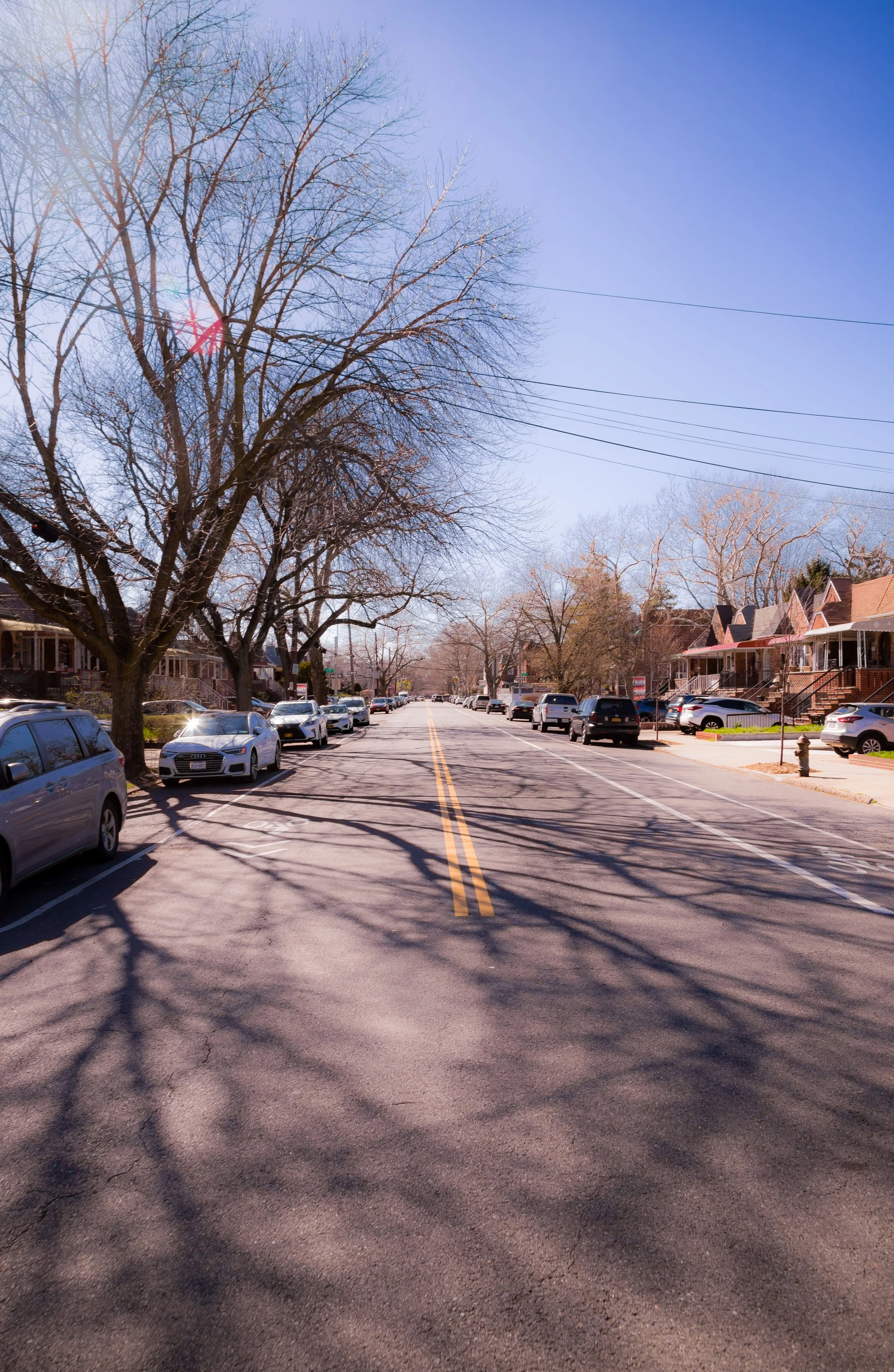 A quiet residential street lined with parked cars on both sides, leafless trees casting shadows on the pavement, and row houses with porches under a clear blue sky.