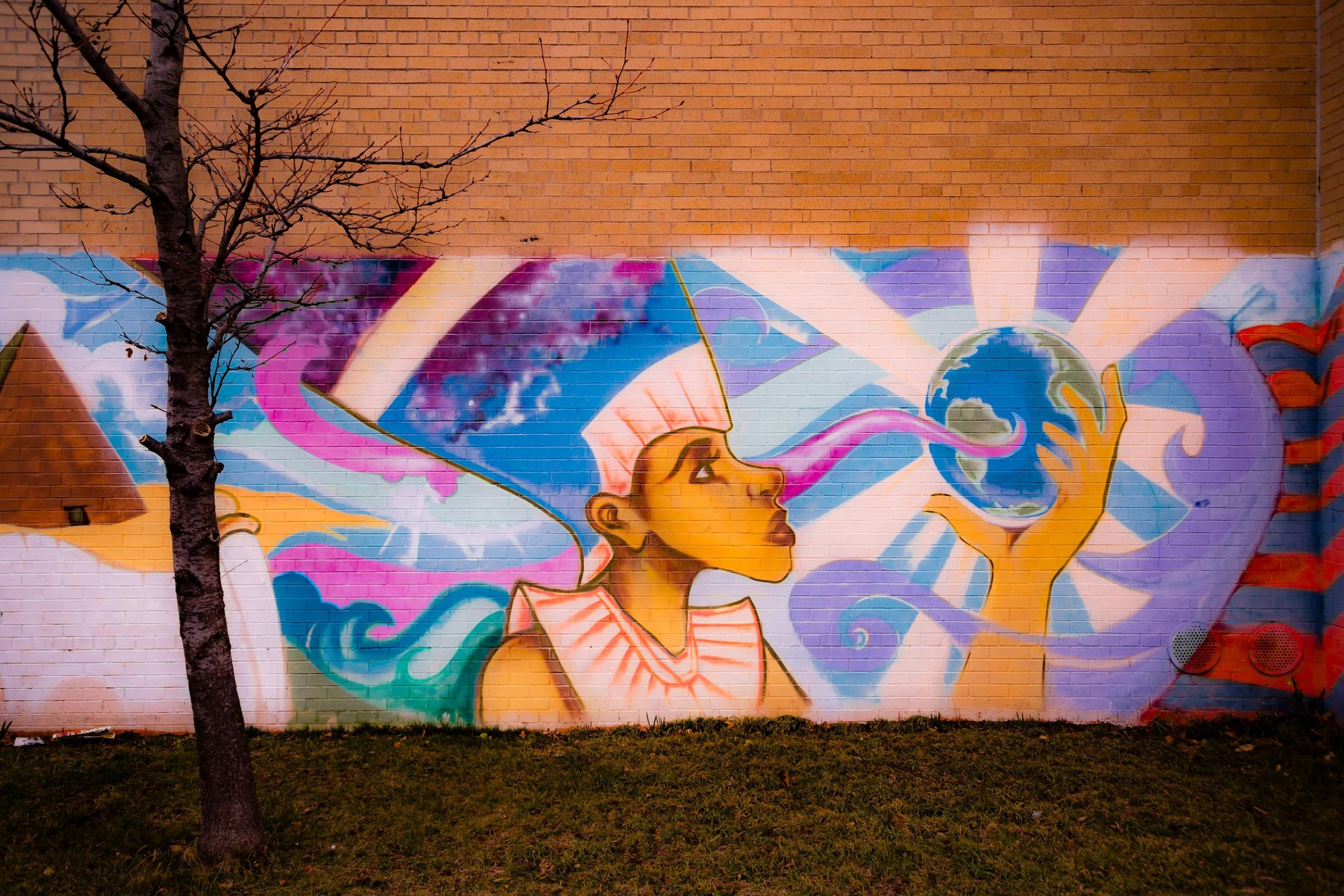 Colorful mural of a woman with a scarf on her head, holding the Earth, surrounded by vibrant abstract shapes and patterns, painted on a brick wall.