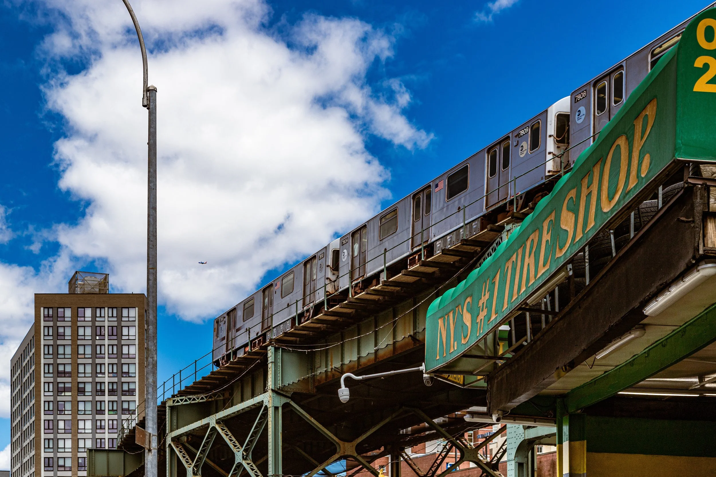 Elevated train on a city track with a blue sky and scattered clouds, a tall building in the background, and green signage for a marketplace.