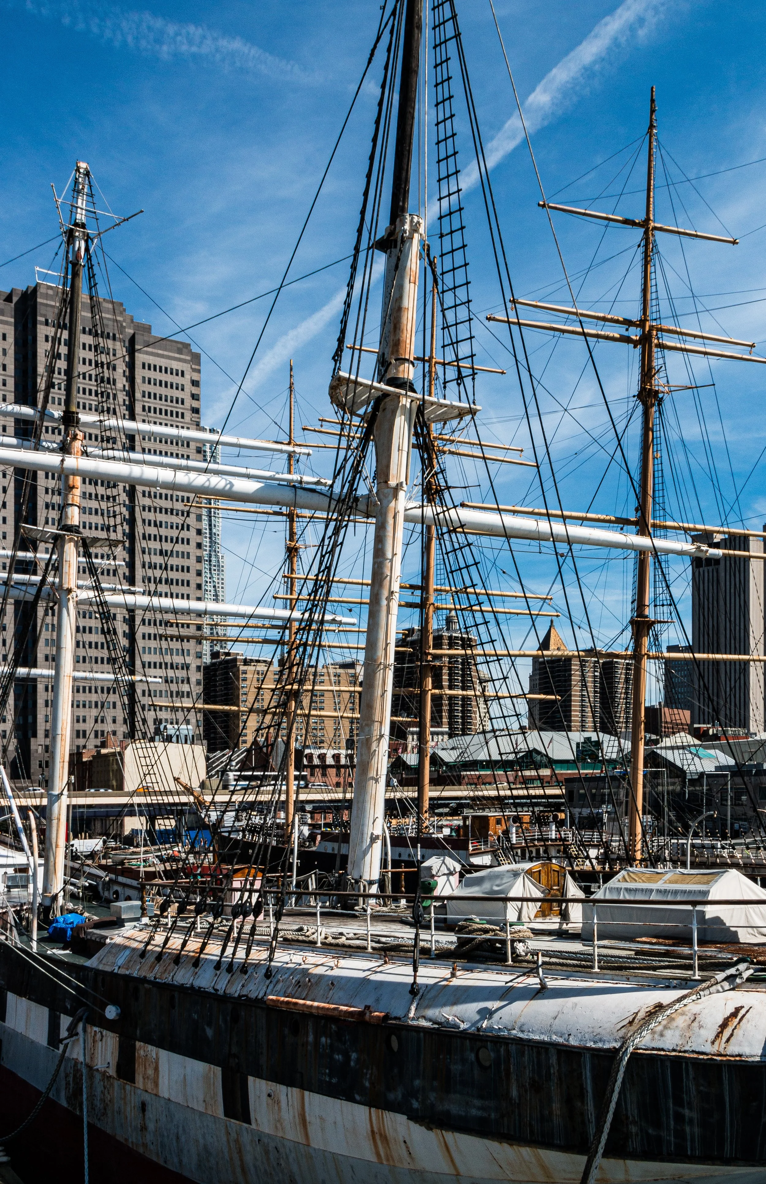 Old, rusty sailing ship docked at a modern marina with tall city buildings in the background, under a partly cloudy blue sky.