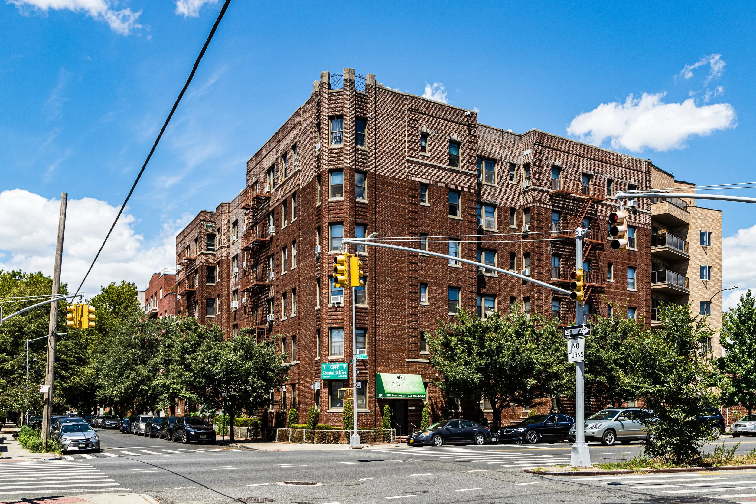 A multi-story brick apartment building with fire escapes on the exterior, situated at a street corner under a blue sky with some clouds. Cars are parked along the street, and trees line the sidewalk.