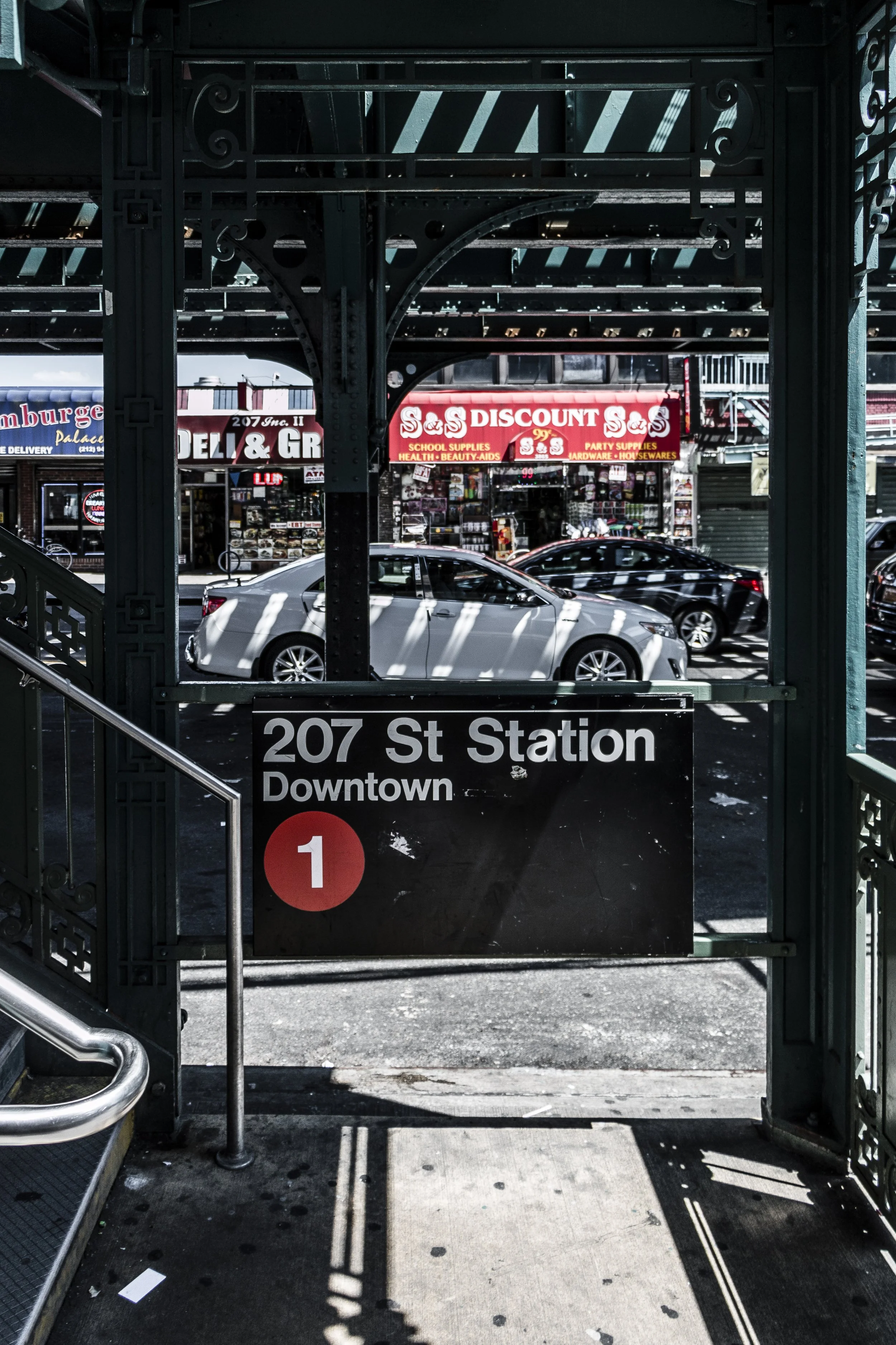 View of a city street through an underground subway exit with a sign indicating '207 St Station, Downtown' and a large red circle with the number 1, cars parked along the street, and storefronts including a discount store and a deli.