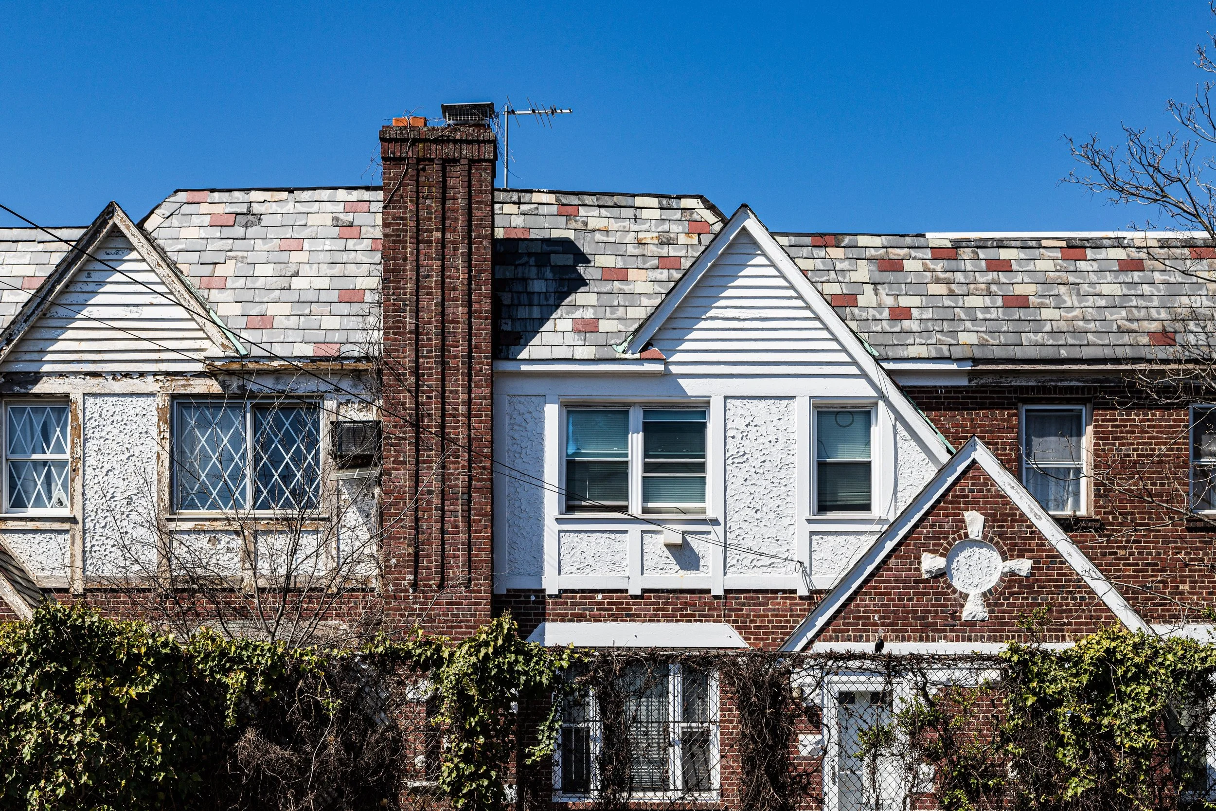A row of attached houses with brick and white exterior, steeply sloped roofs with multicolored shingles, a prominent brick chimney, and a blue sky in the background, with some leafless trees and vines in the foreground.