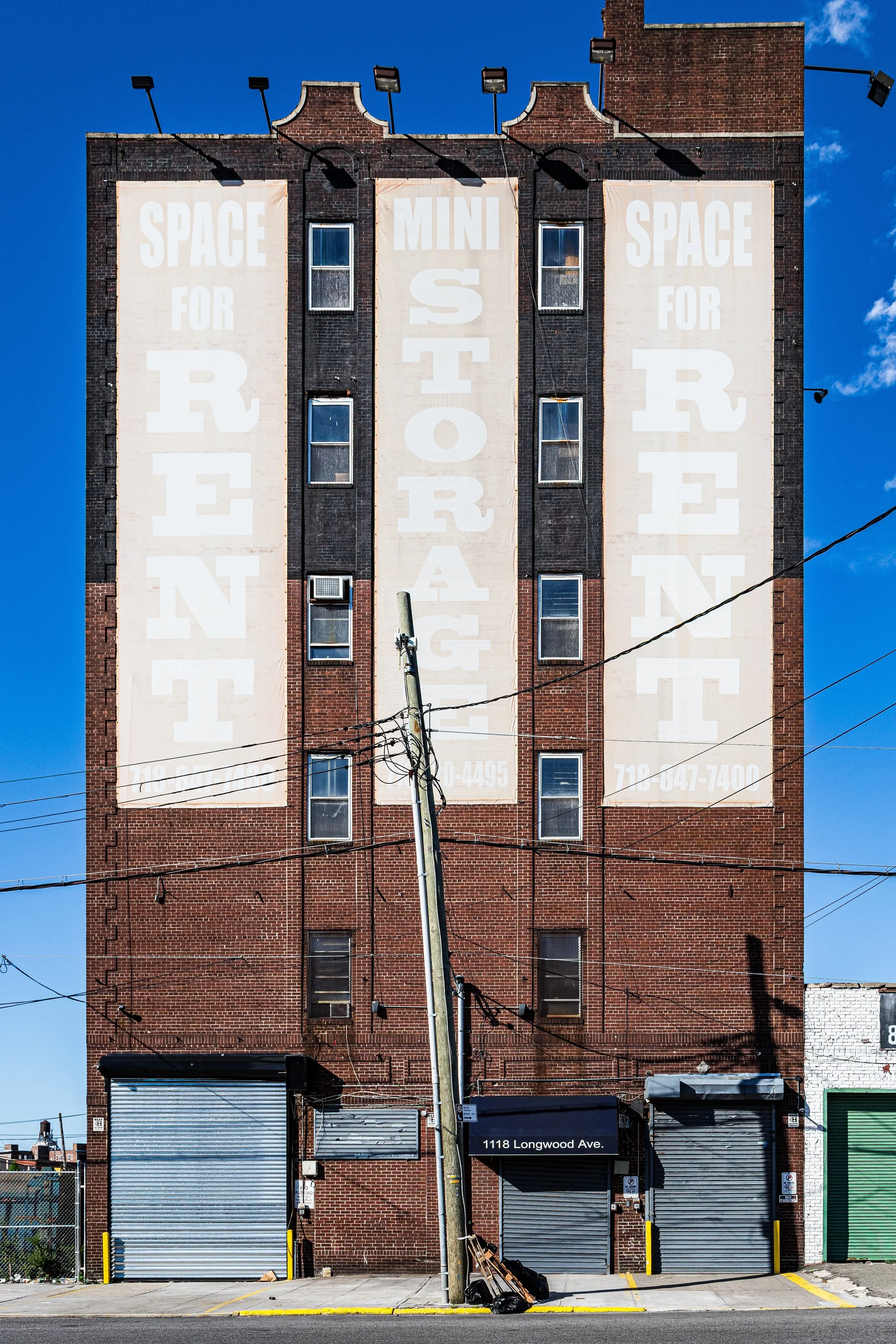 The side of a tall brick building with large painted signs that promote space rental, including phrases like 'Space for Rent' and 'Mini Storage.' The building has several windows, some with air conditioning units, and two rolling metal garage doors at the bottom. A street sign indicates "1118 Longwood Ave." In front of the building, a tilted utility pole with wires extends across the scene, against a bright blue sky.