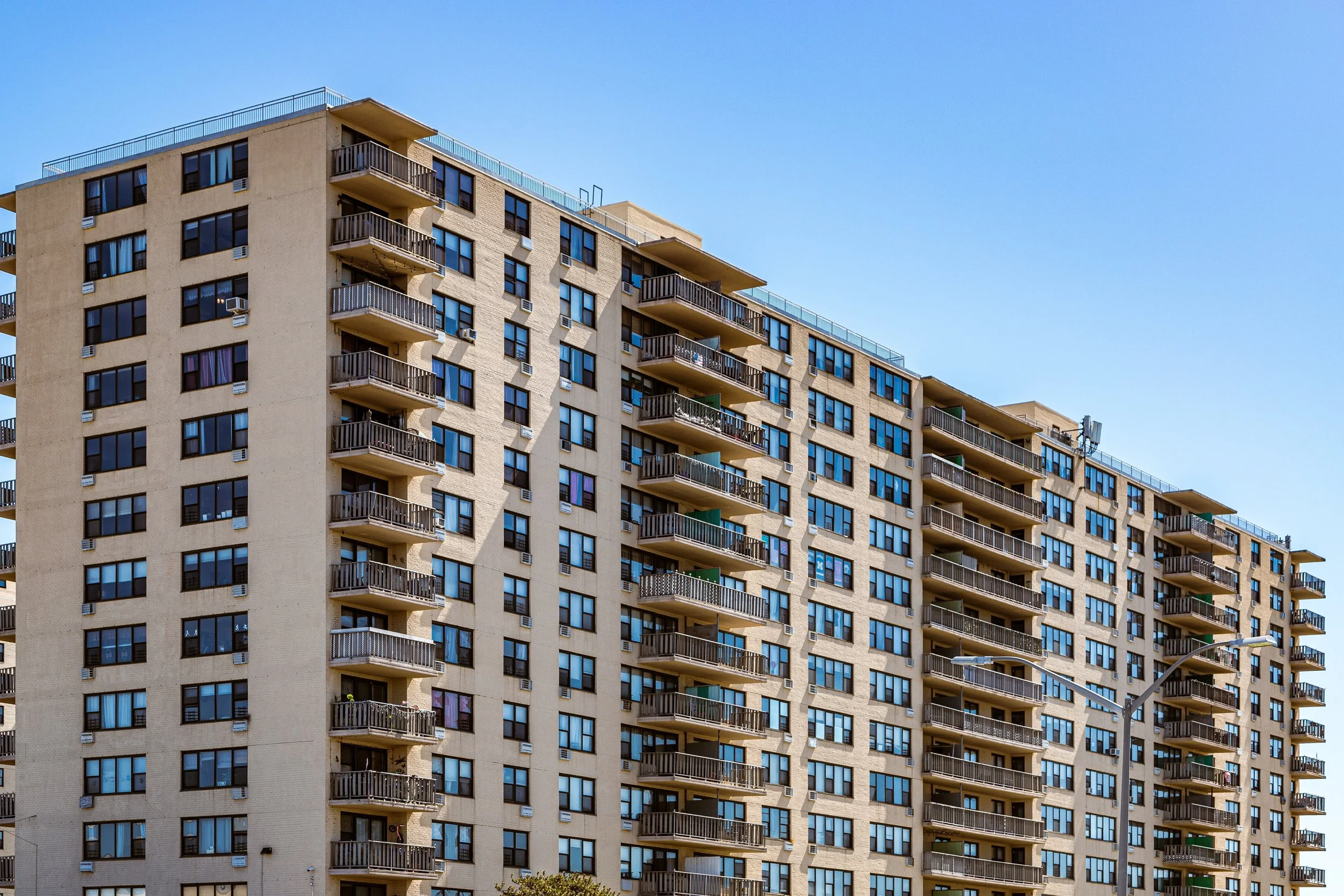 A large beige apartment building with multiple floors and balconies, set against a clear blue sky.