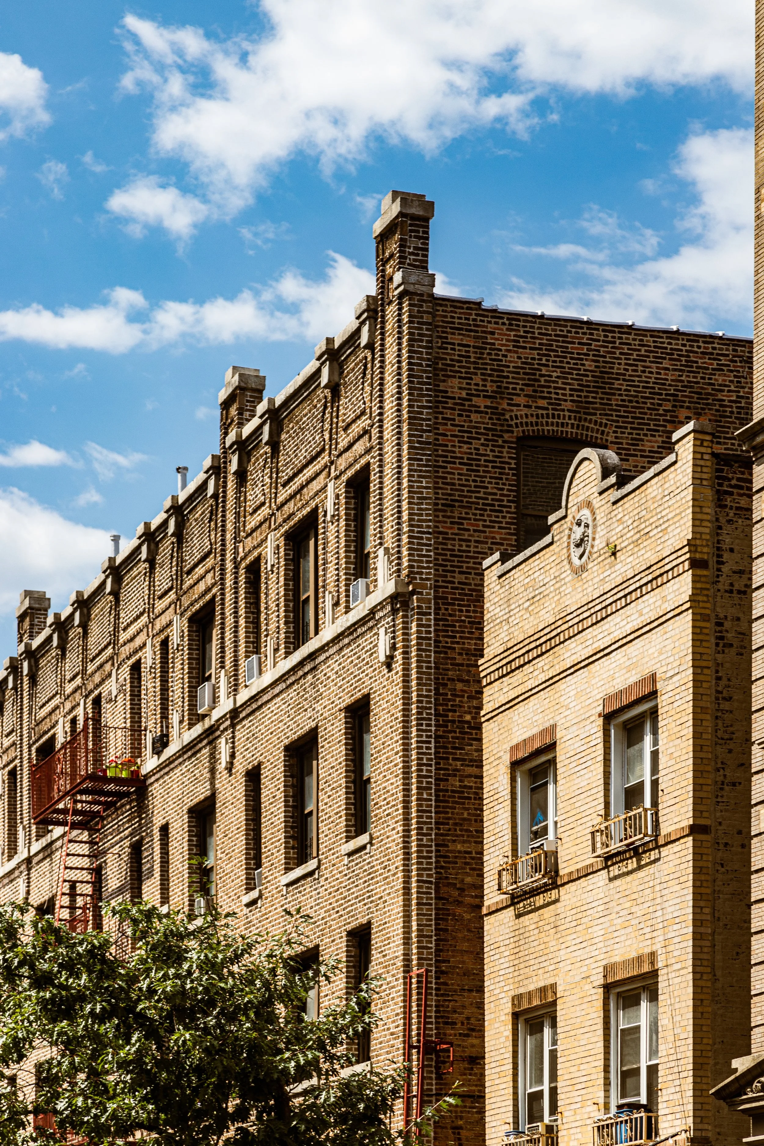 Close-up view of two tan and brown brick apartment buildings with fire escapes and small balconies, set against a bright blue sky with scattered clouds.