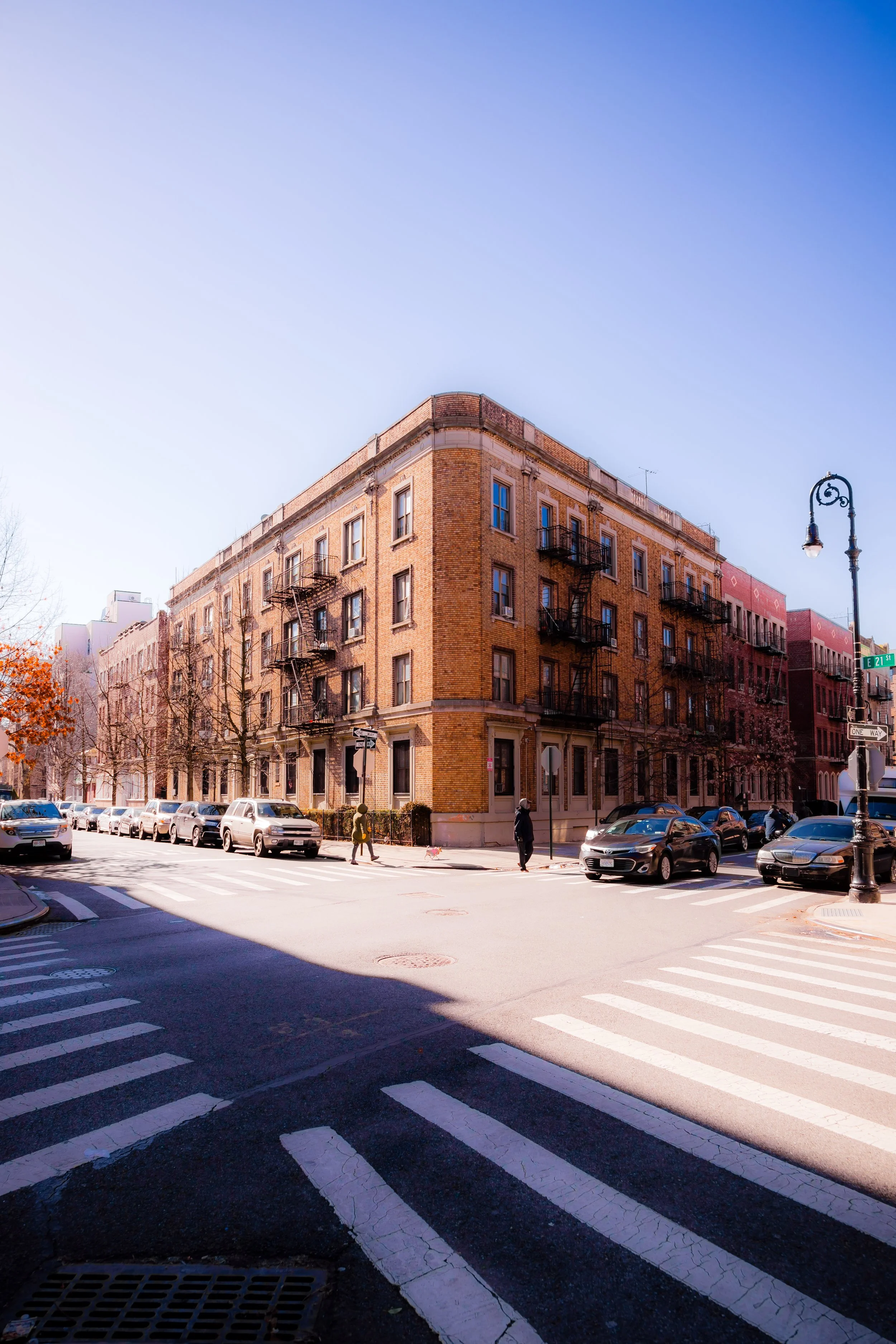 A street corner in an urban neighborhood with a large brick building, parked cars, pedestrians, street signs, and a vintage street lamp, under a clear blue sky.
