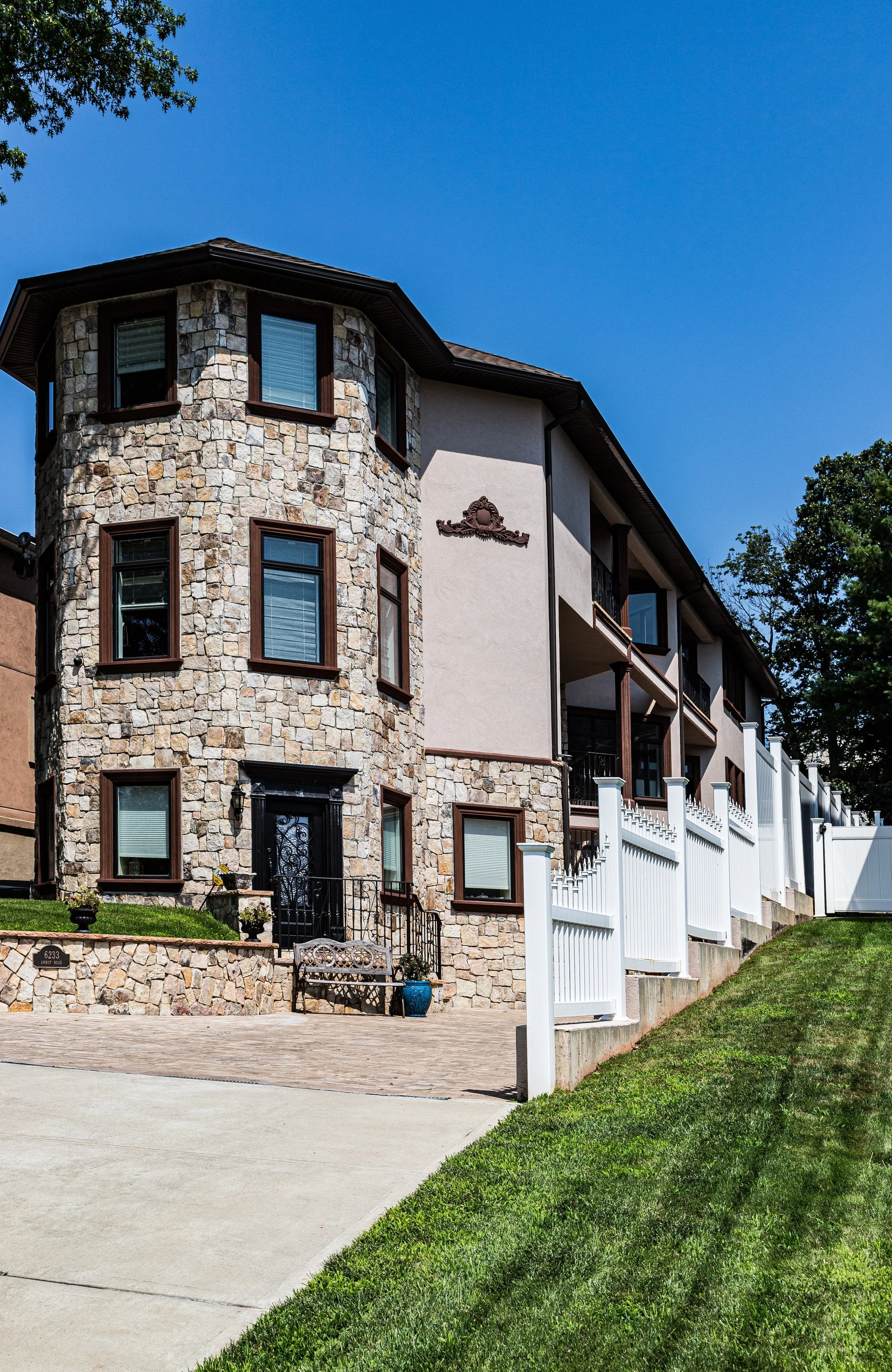 Modern residential building with a stone facade and multiple balconies, white fence, and a paved driveway under a clear blue sky.