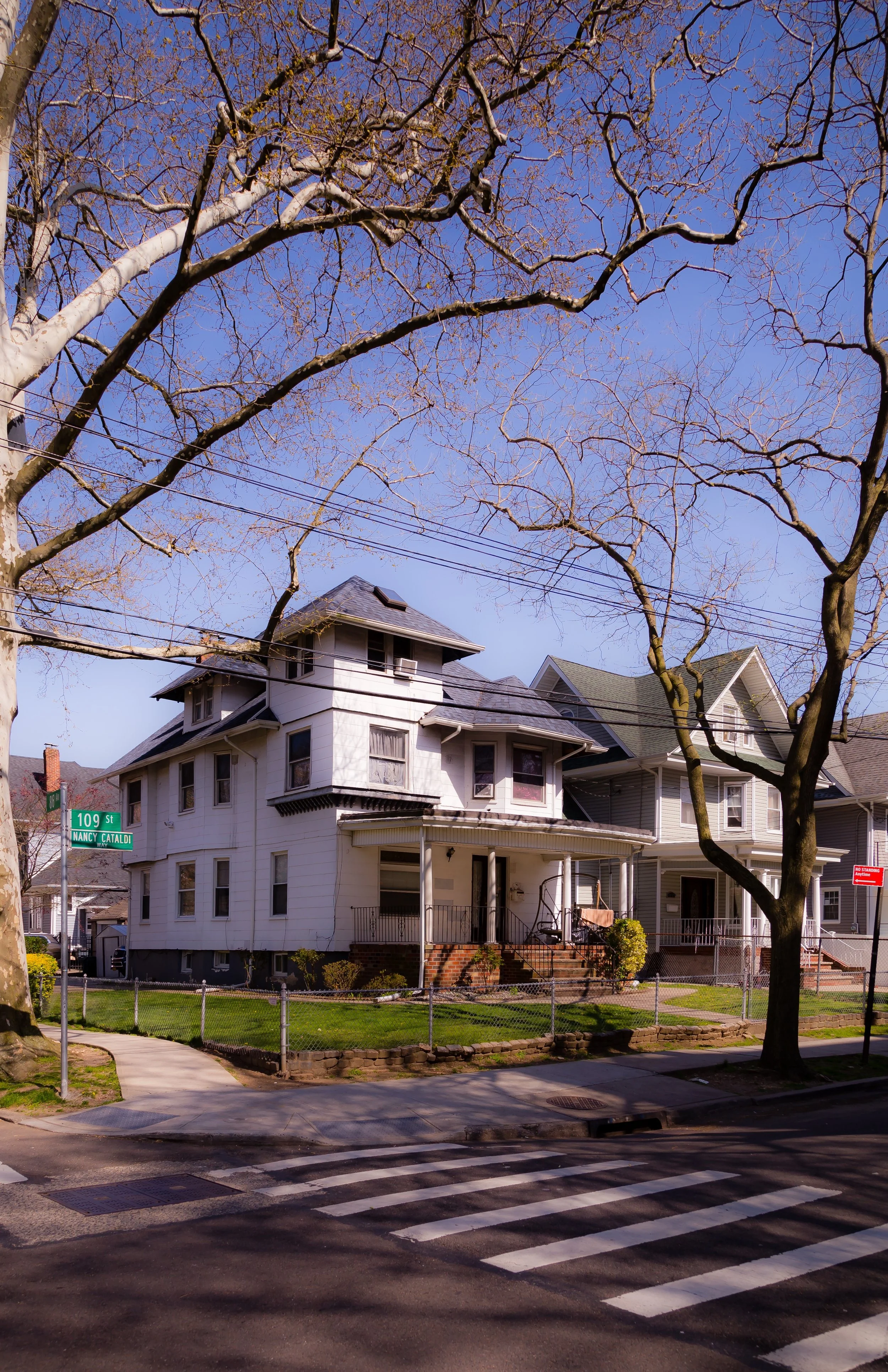 A street corner with a white, multi-story house and a smaller house behind it, leafless trees, electric wires, and a pedestrian crosswalk.