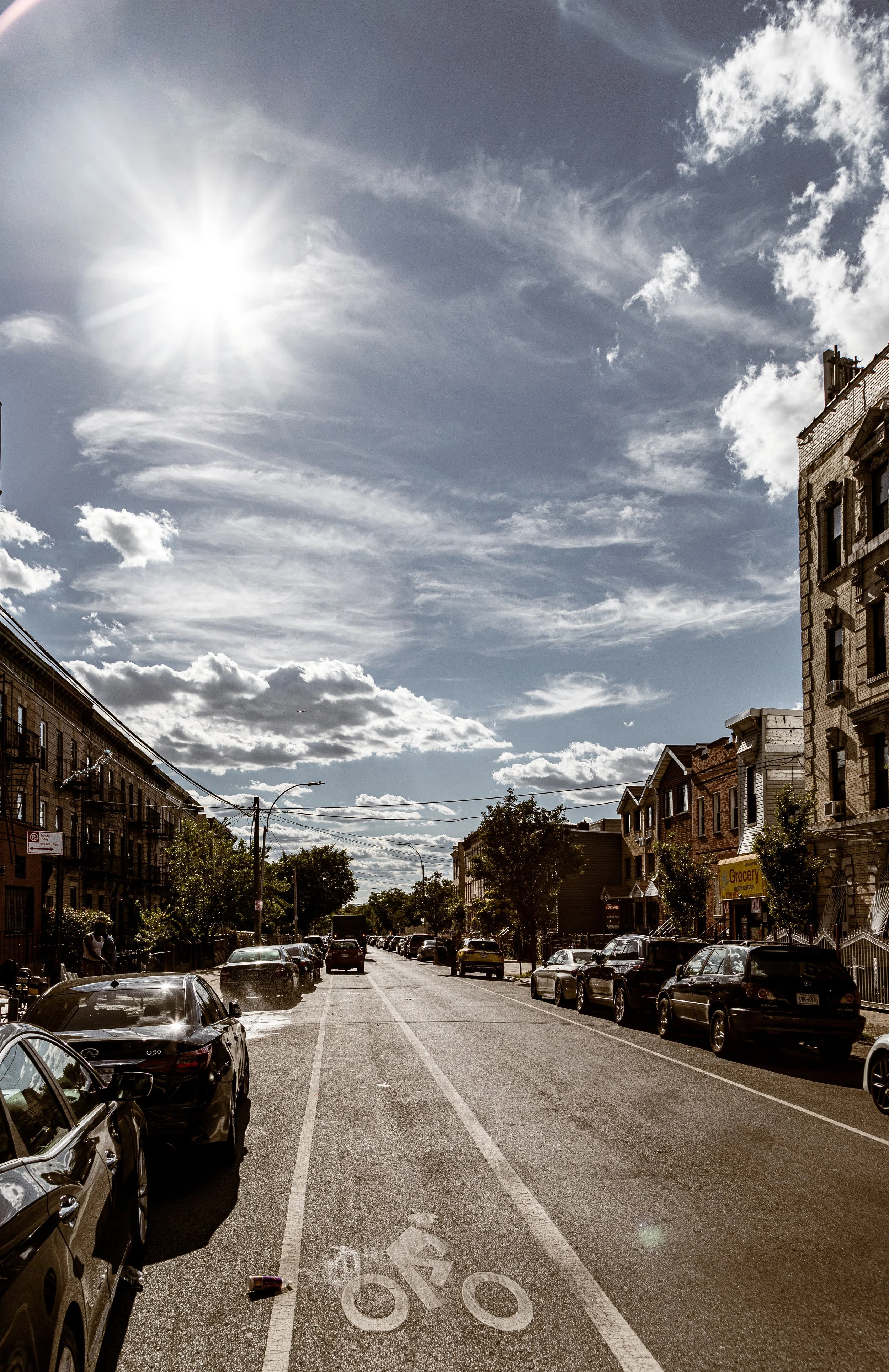 A sunny urban street with parked cars on both sides, trees lining the sidewalks, residential buildings, and a bright sky with clouds.