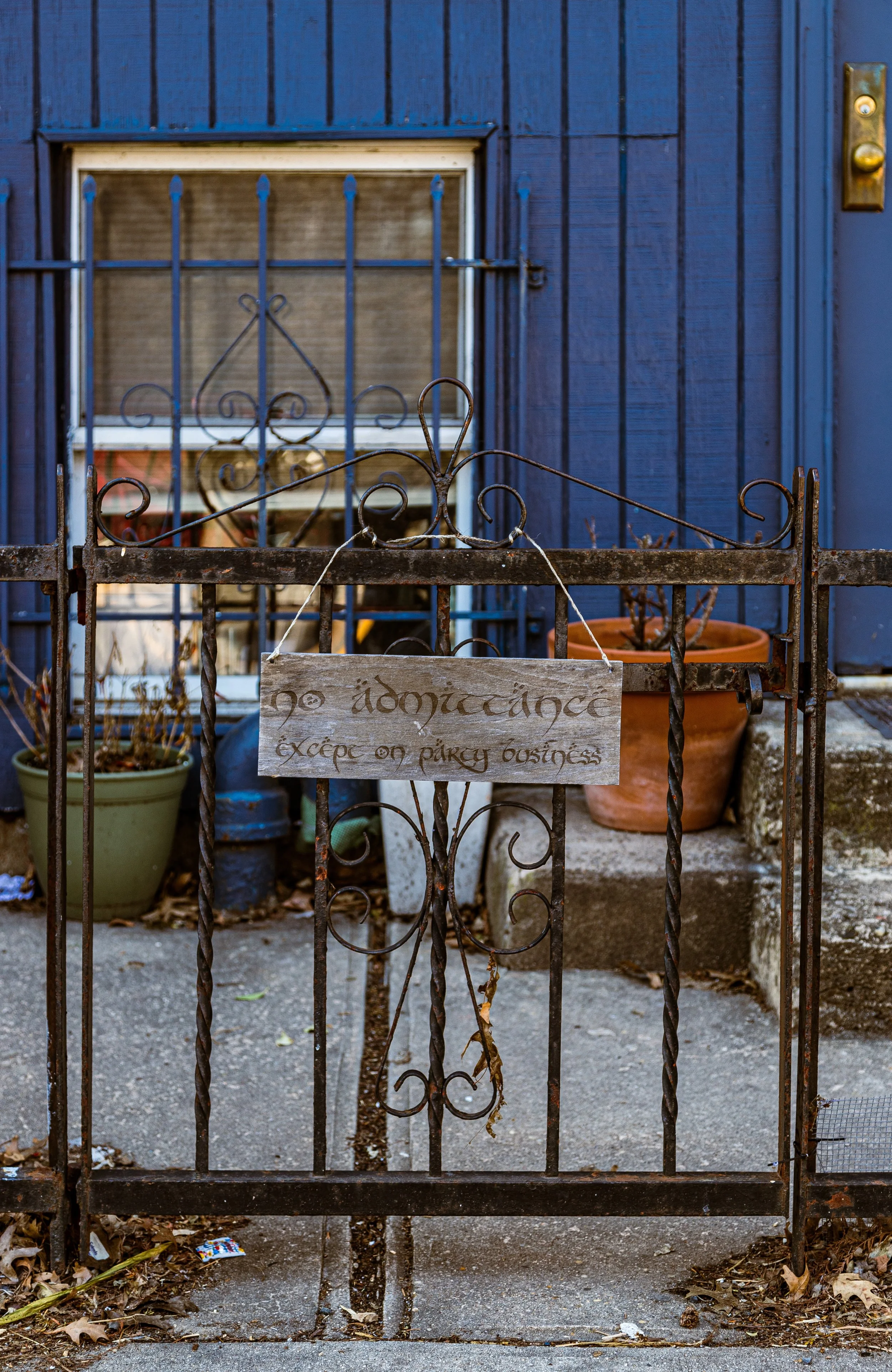 A wrought iron gate with a sign that reads 'No admittance except on party business' in front of a blue house with a window and several potted plants.