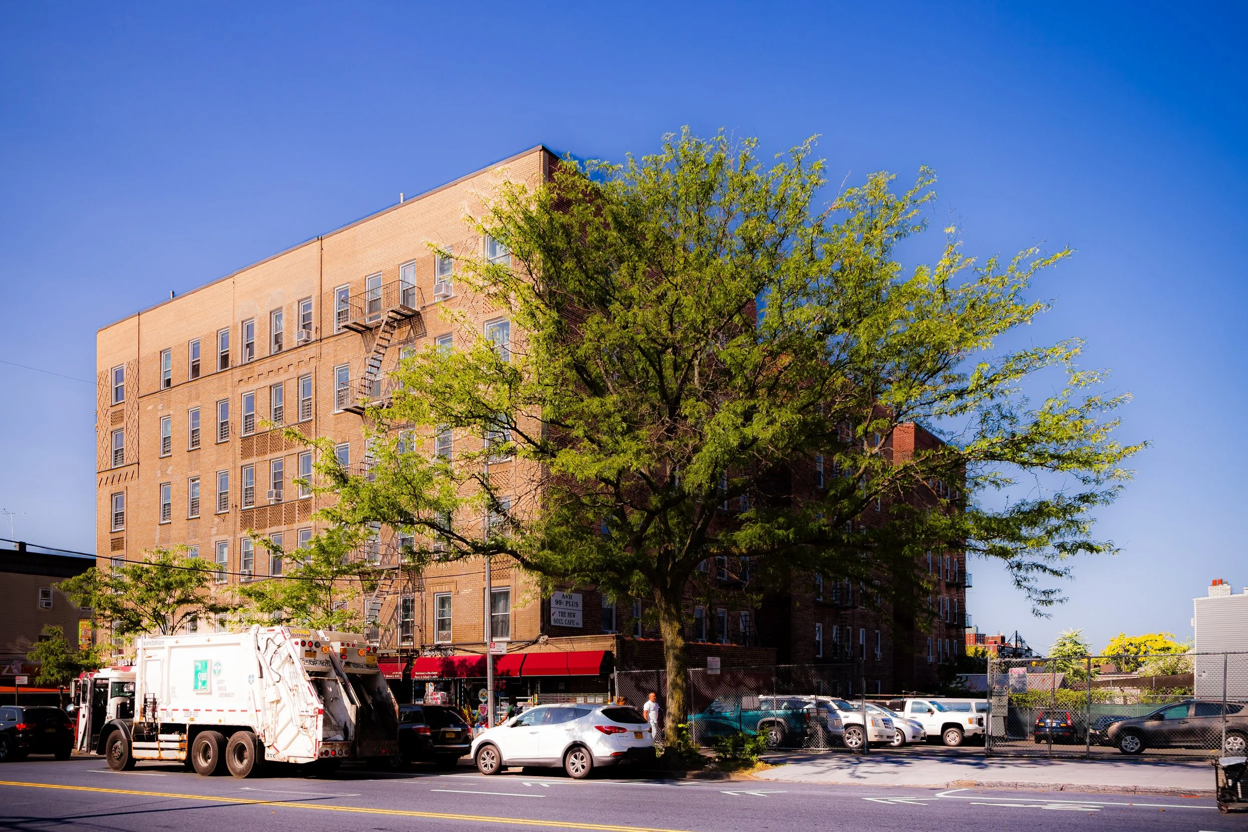 A brick apartment building with a fire escape on the side, a large green tree in front, and a parking lot with cars, a garbage truck, and pedestrians on the street under a clear blue sky.