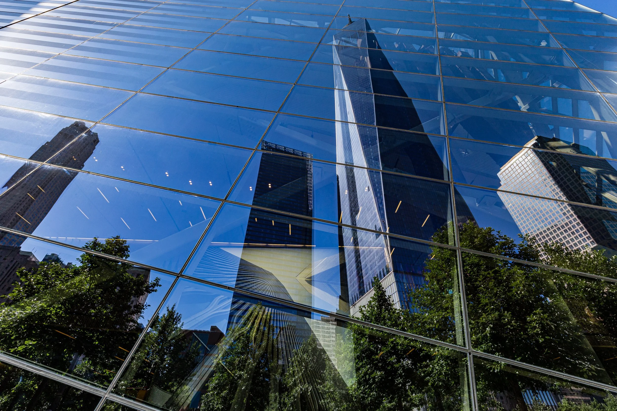 Sky reflected on the glass windows of a tall skyscraper, showing surrounding buildings and trees.