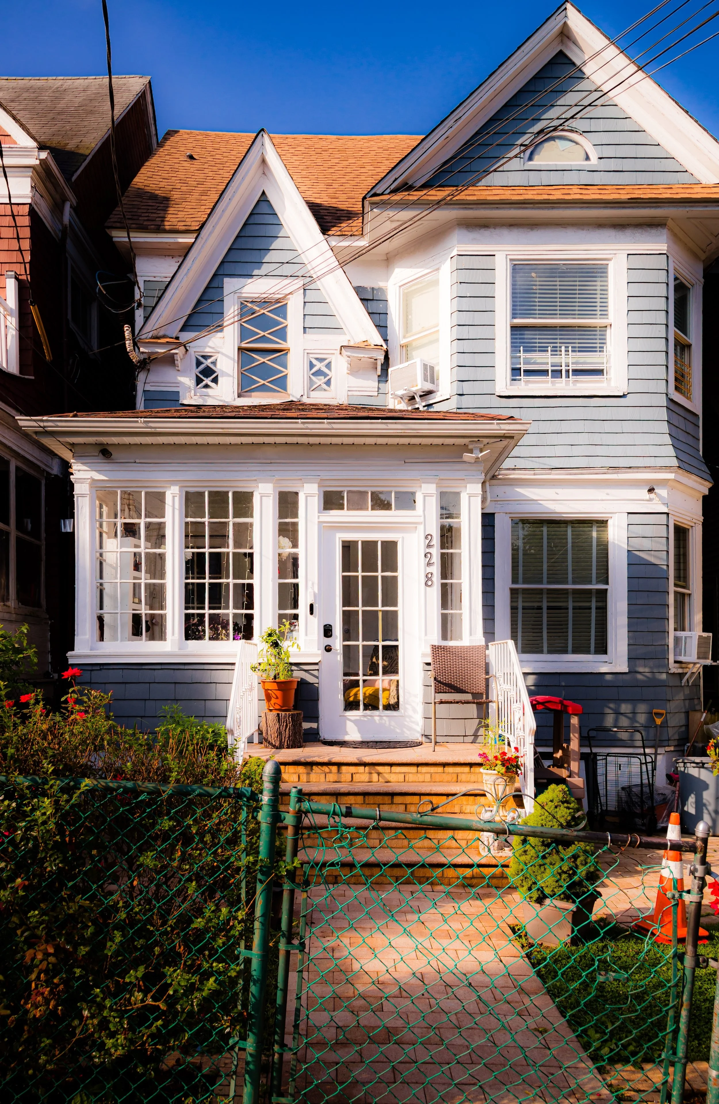 Front view of a blue Victorian house with white trim, large windows, a small porch with steps, potted plants, and a garden in front, enclosed by a green metal fence.