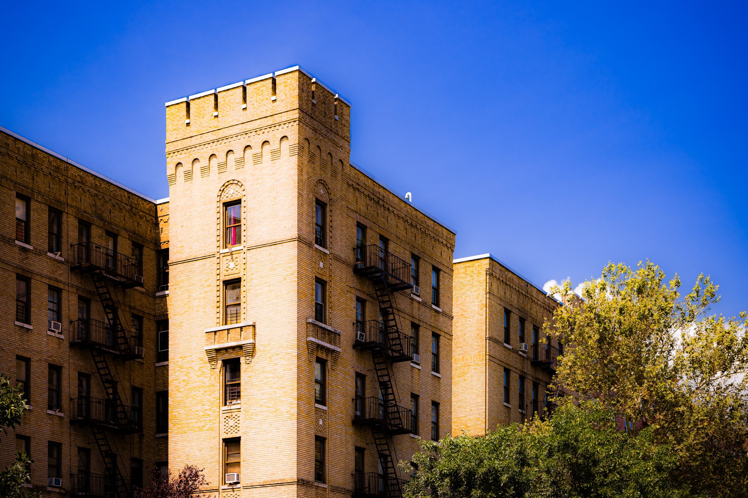 A brick apartment building with fire escapes, a tower-like structure, and trees in front, under a clear blue sky.