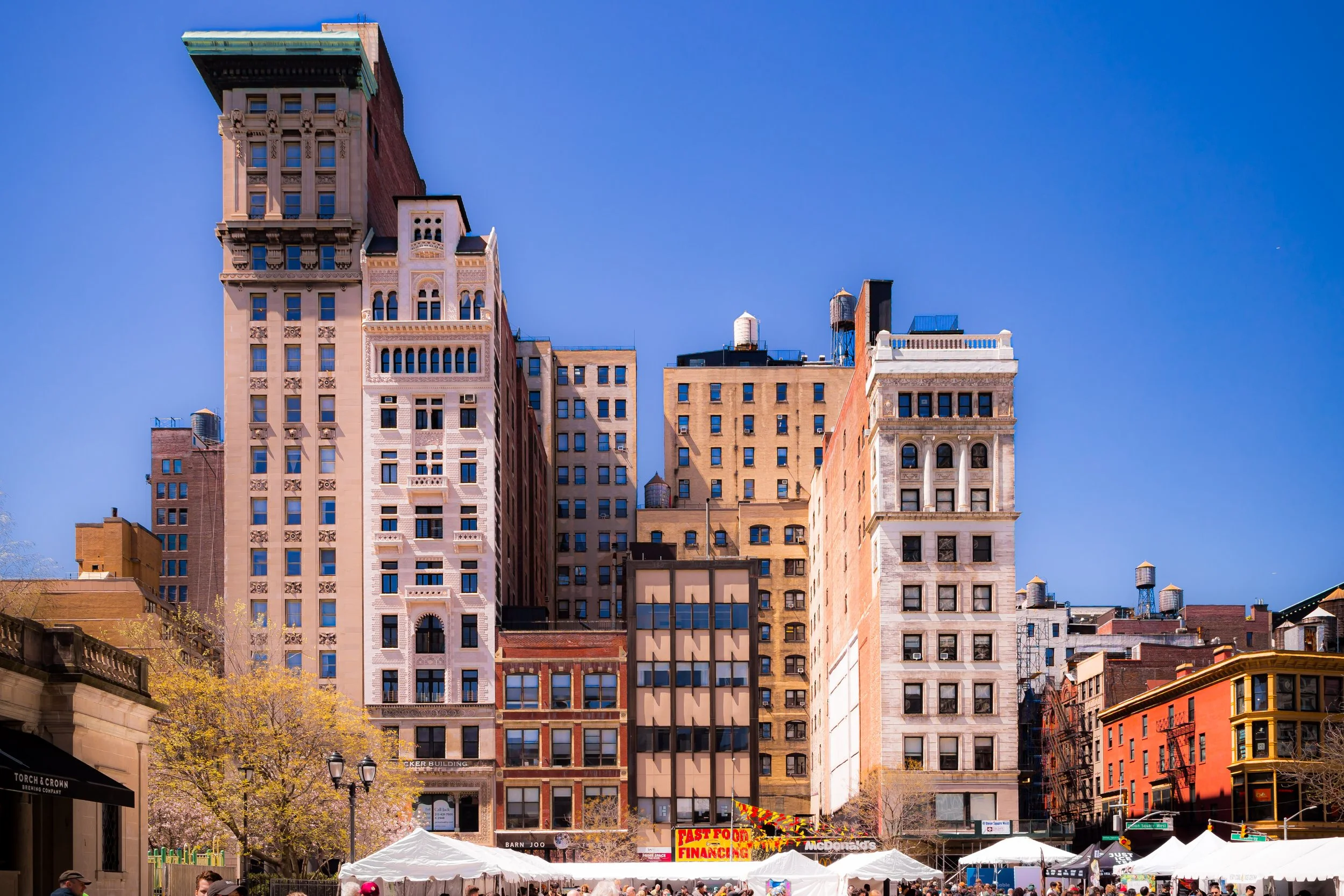 Multiple high-rise buildings in New York City with tents and stalls in the foreground, under a blue sky.