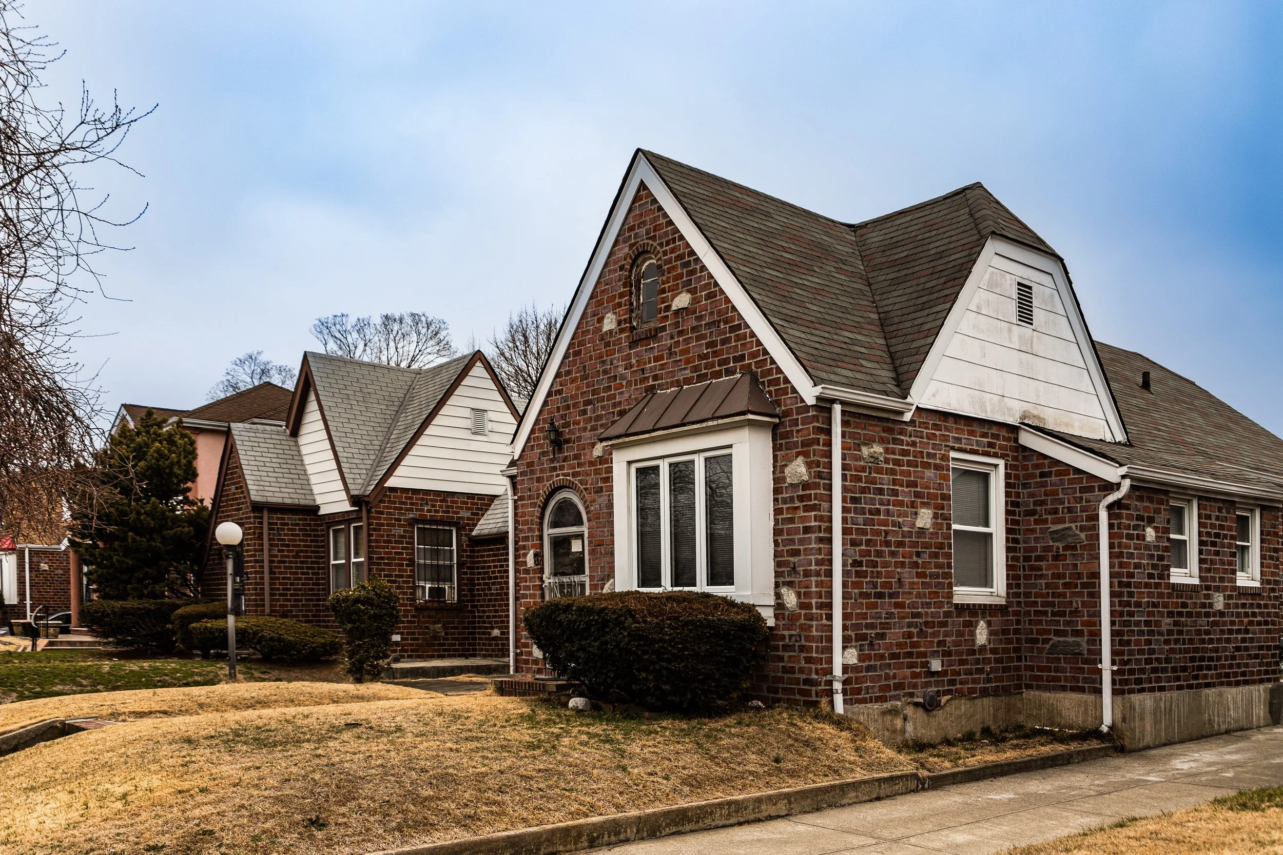 A brick house with a steep roof, white trim, and multiple windows on a suburban street with a lawn and sidewalk