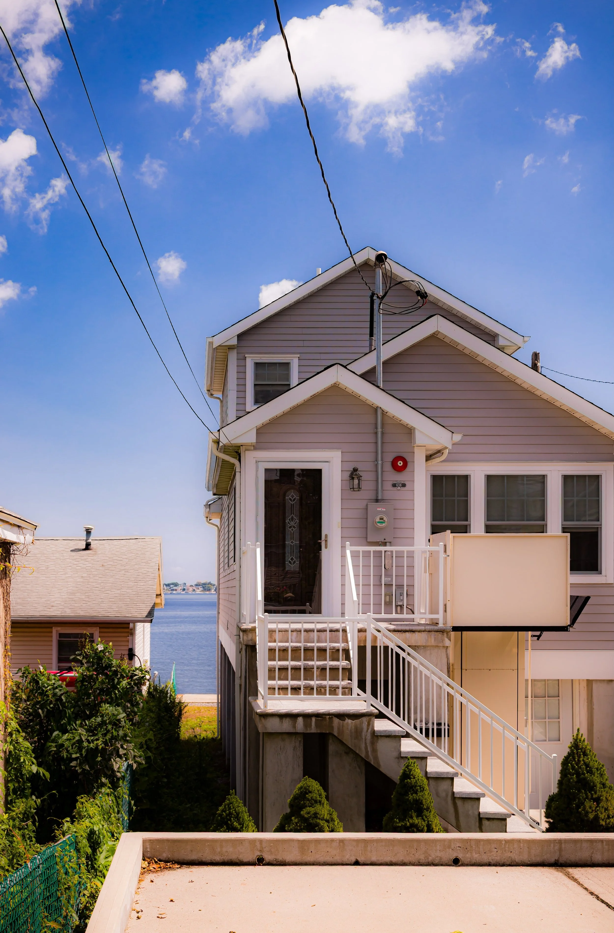 A pink house on stilts with a staircase leading up to the front door, with water and a clear blue sky in the background.