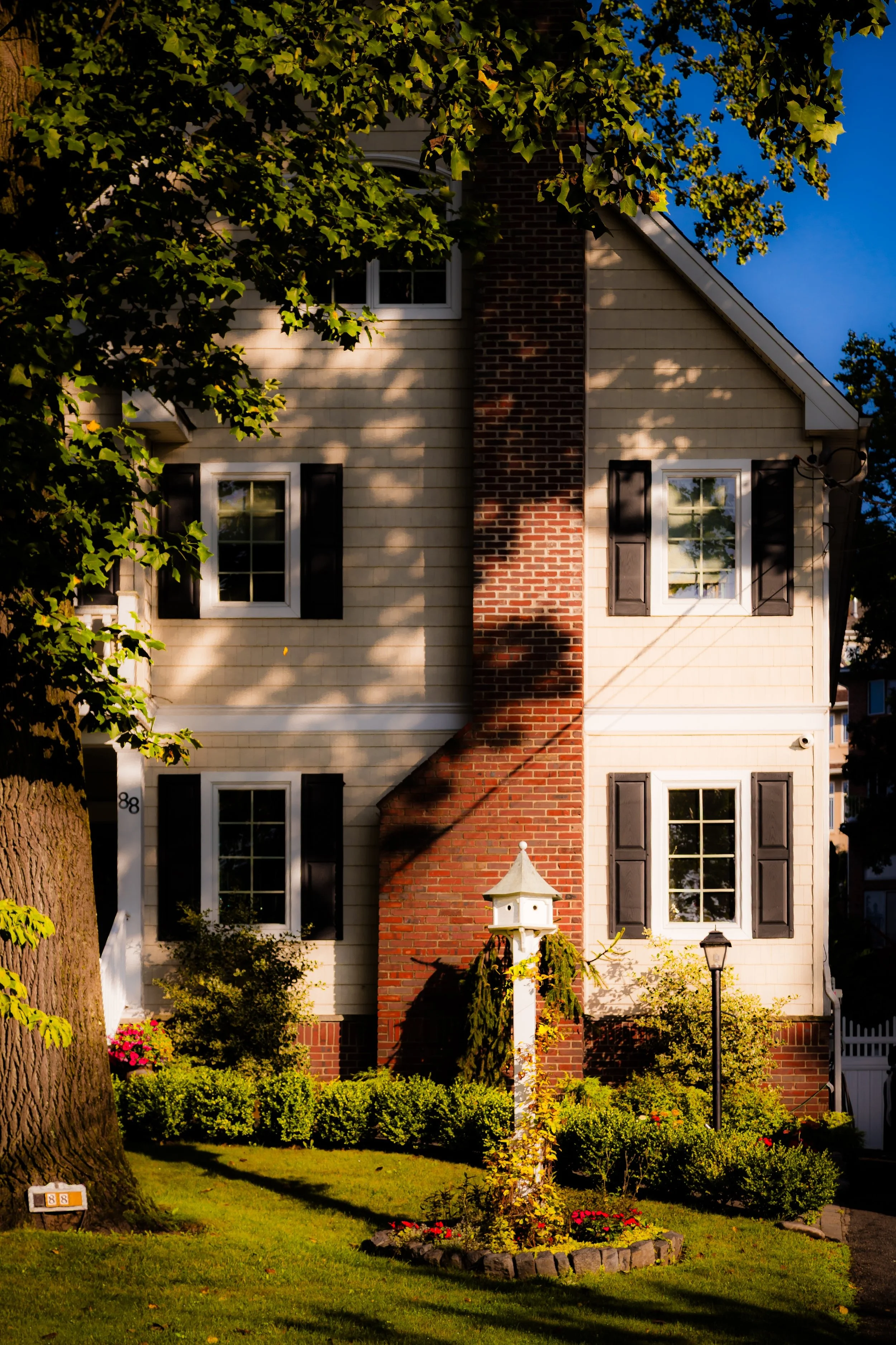 A two-story house with beige siding, black shutters, and a large brick chimney. The front yard has green grass, bushes, and a small flower bed with red and pink flowers. There is a white birdhouse on a post, a lamp post, and large leafy trees casting shadows.