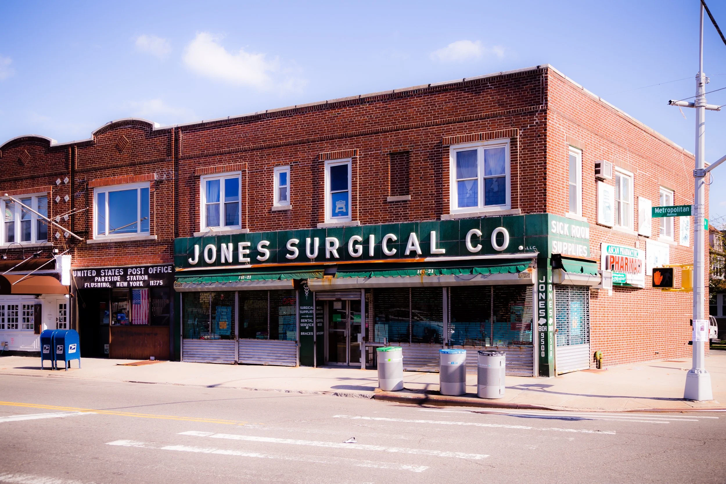 A two-story brick building with a green sign reading 'Jones Surgical Co.' and signs for medical supplies. A USPS mailbox and trash bins are on the sidewalk, with a street and traffic signs visible.