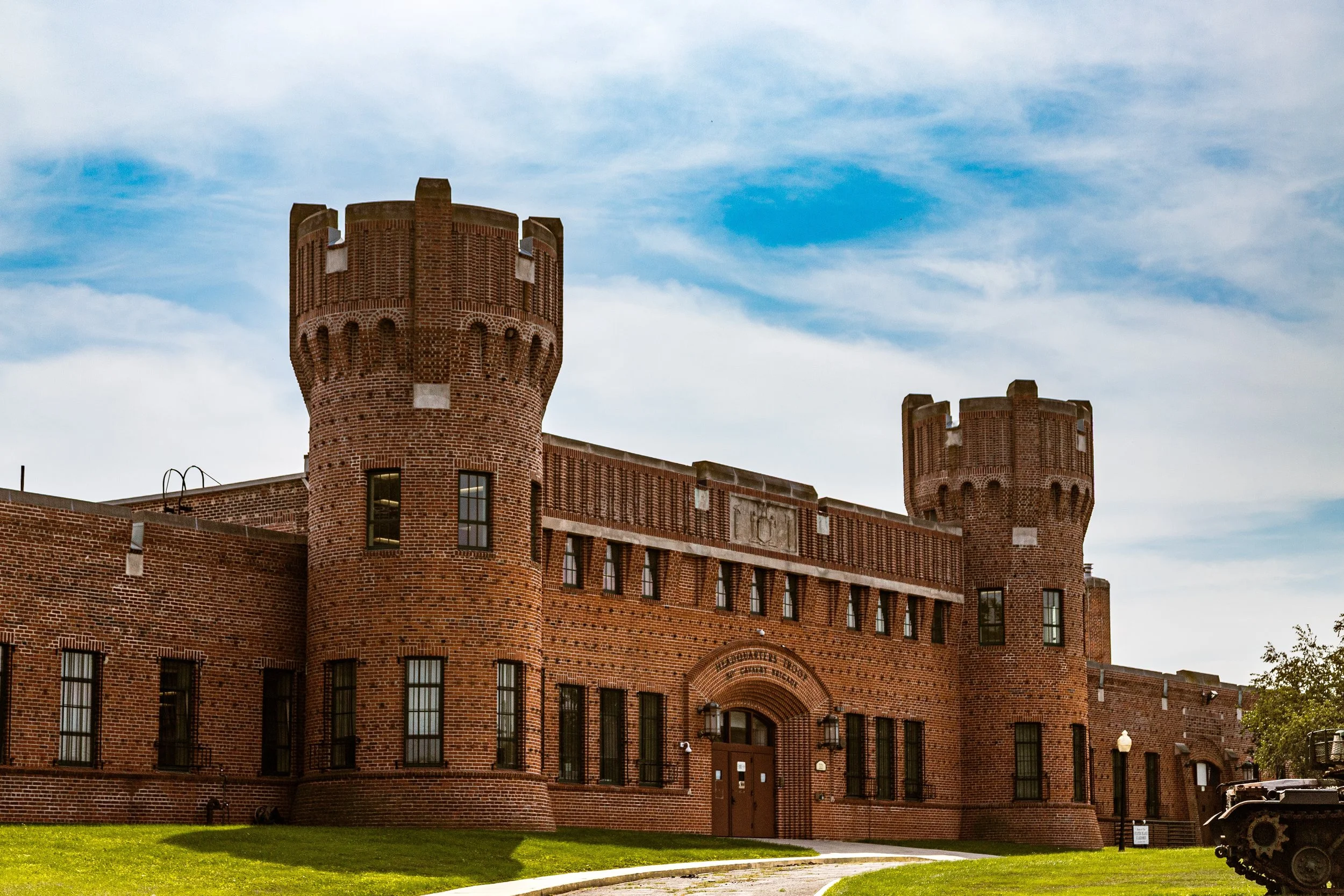 Historic red brick building with two corner towers, arched entrance, and surrounded by green lawn and walking path under a partly cloudy sky.
