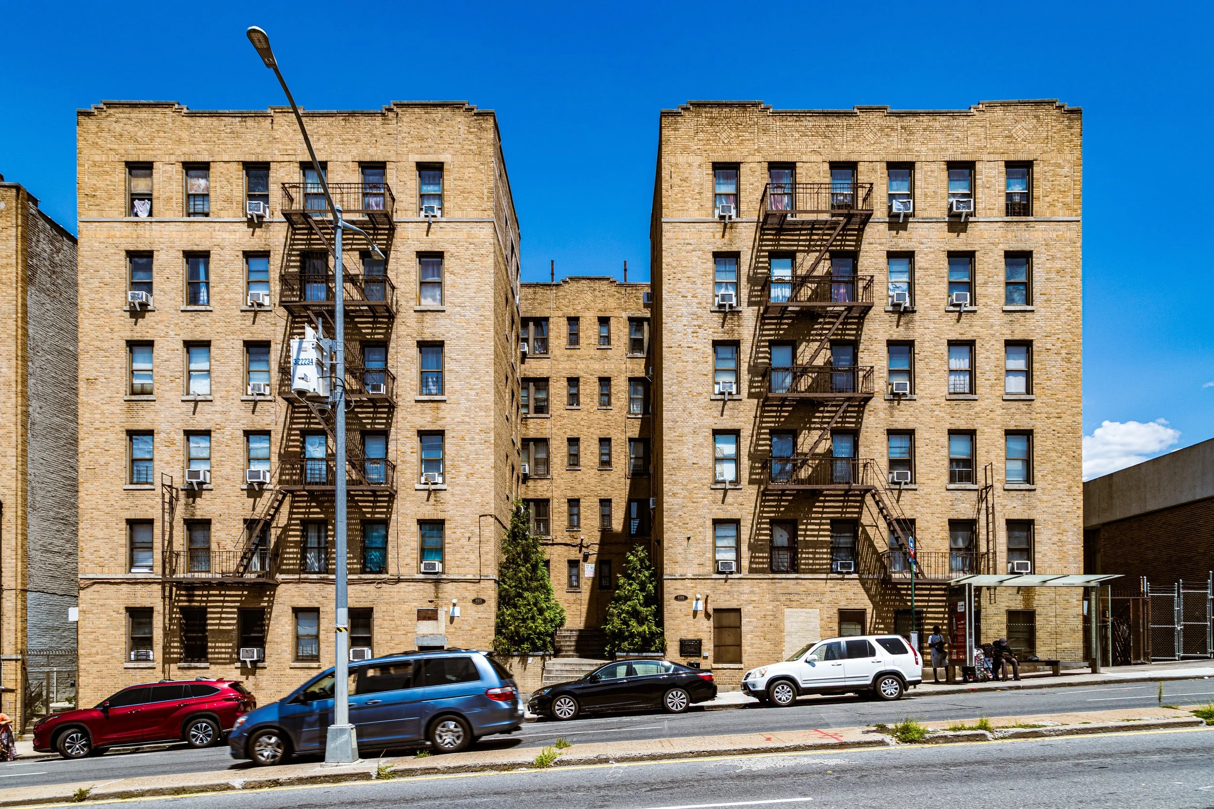 Three brick apartment buildings with fire escapes, parked cars in front, sidewalk and bus stop with people, against a clear blue sky.