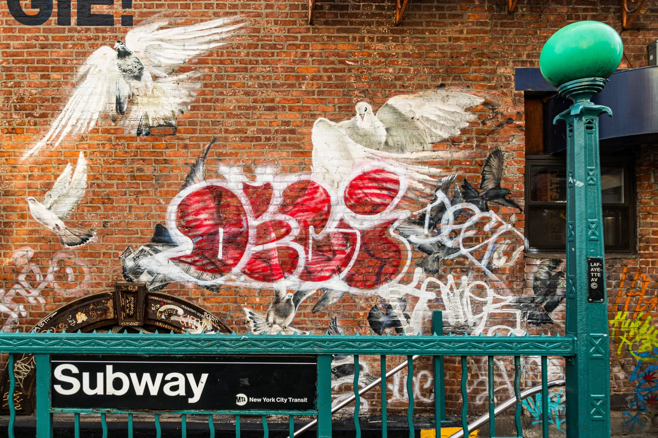 Street art mural on a brick wall featuring a flock of white birds flying and a large red heart with graffiti tags. A green subway entrance with a sign reading 'Subway' and 'New York City Transit' is in the foreground.