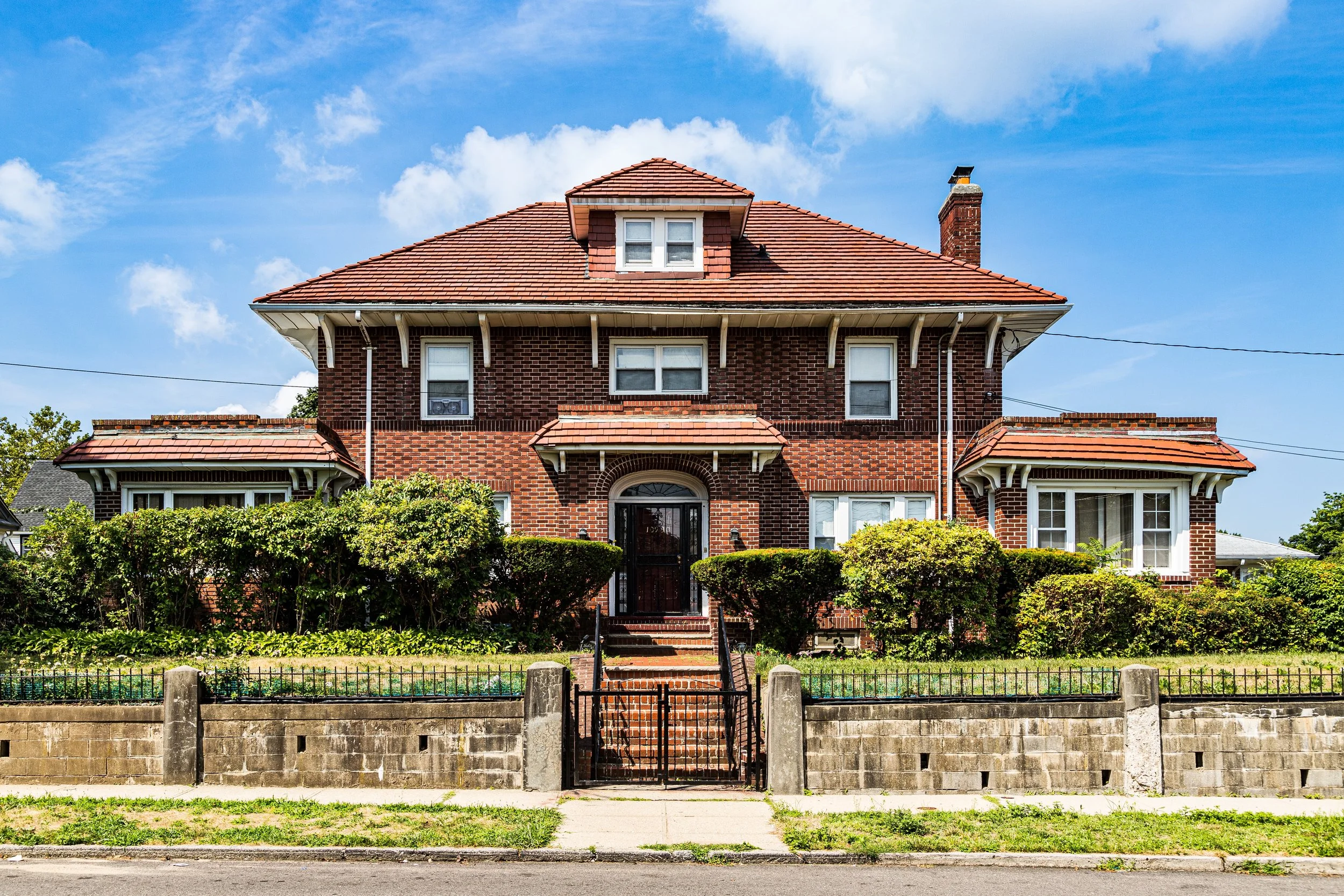 A large red brick house with a red tiled roof, multiple windows, and surrounded by green bushes and a low stone wall with a gate.