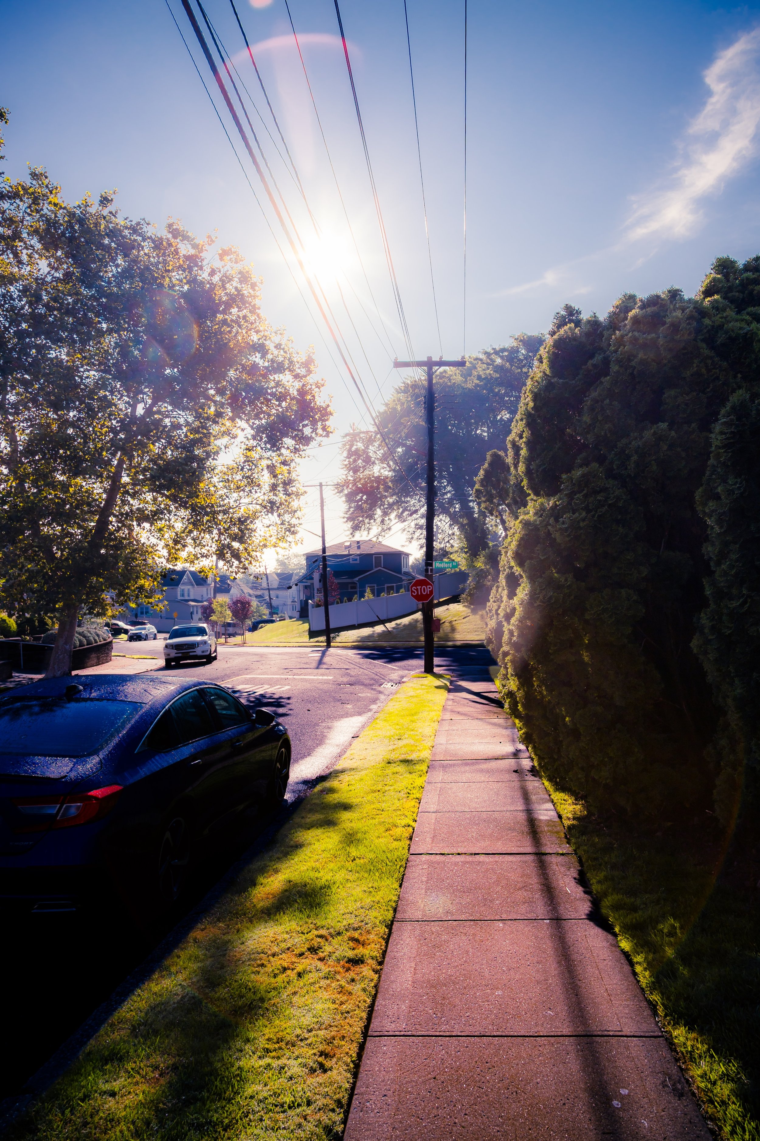 A sunny suburban street scene with a sidewalk, green grass, parked cars, trees, and a stop sign at an intersection. Power lines run overhead, with the sun shining brightly in a clear blue sky.