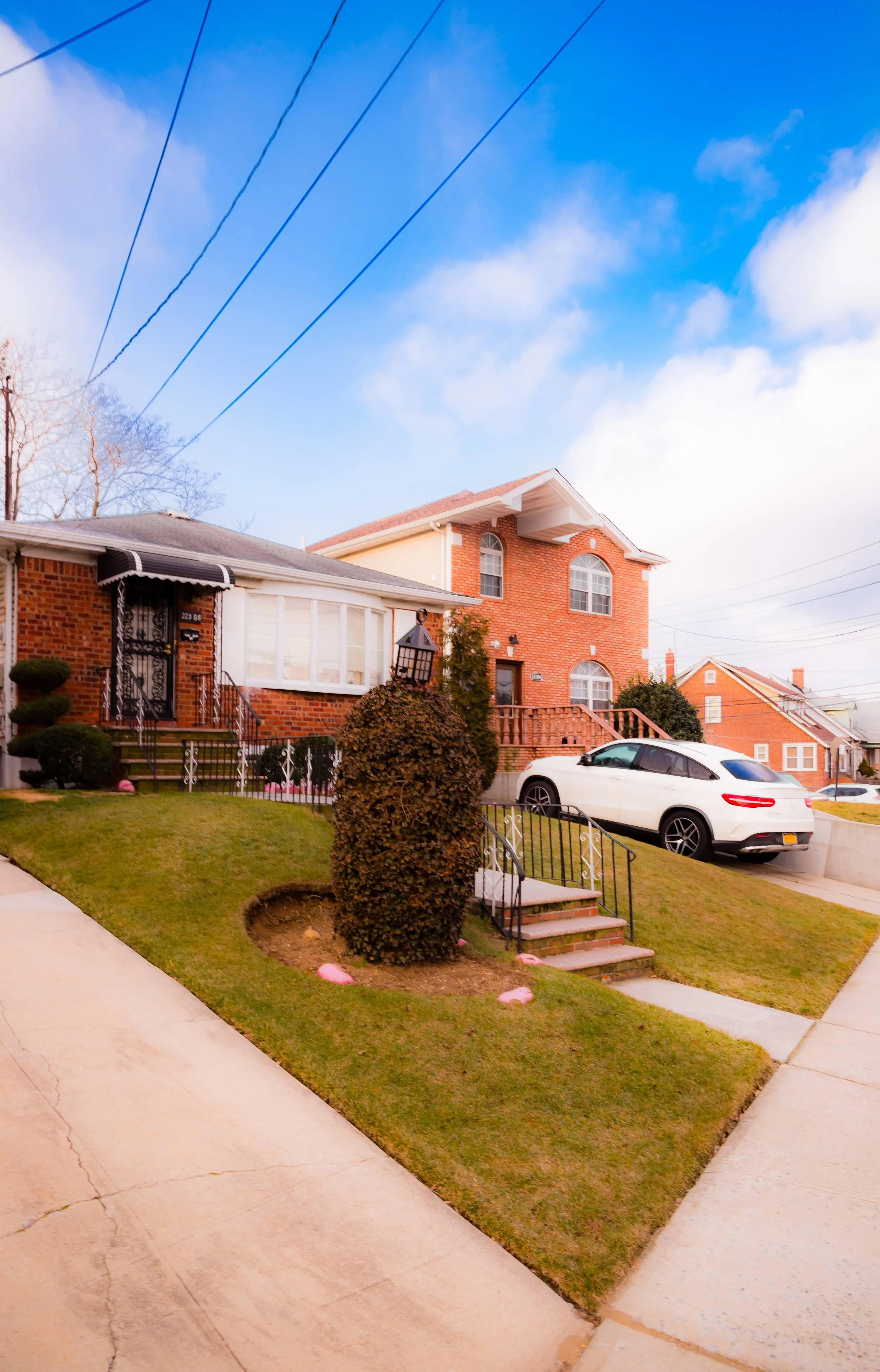 A residential neighborhood with a brick house featuring a front porch, a small staircase, a manicured lawn with a shrub, and a white car parked on an incline, under a partly cloudy blue sky.