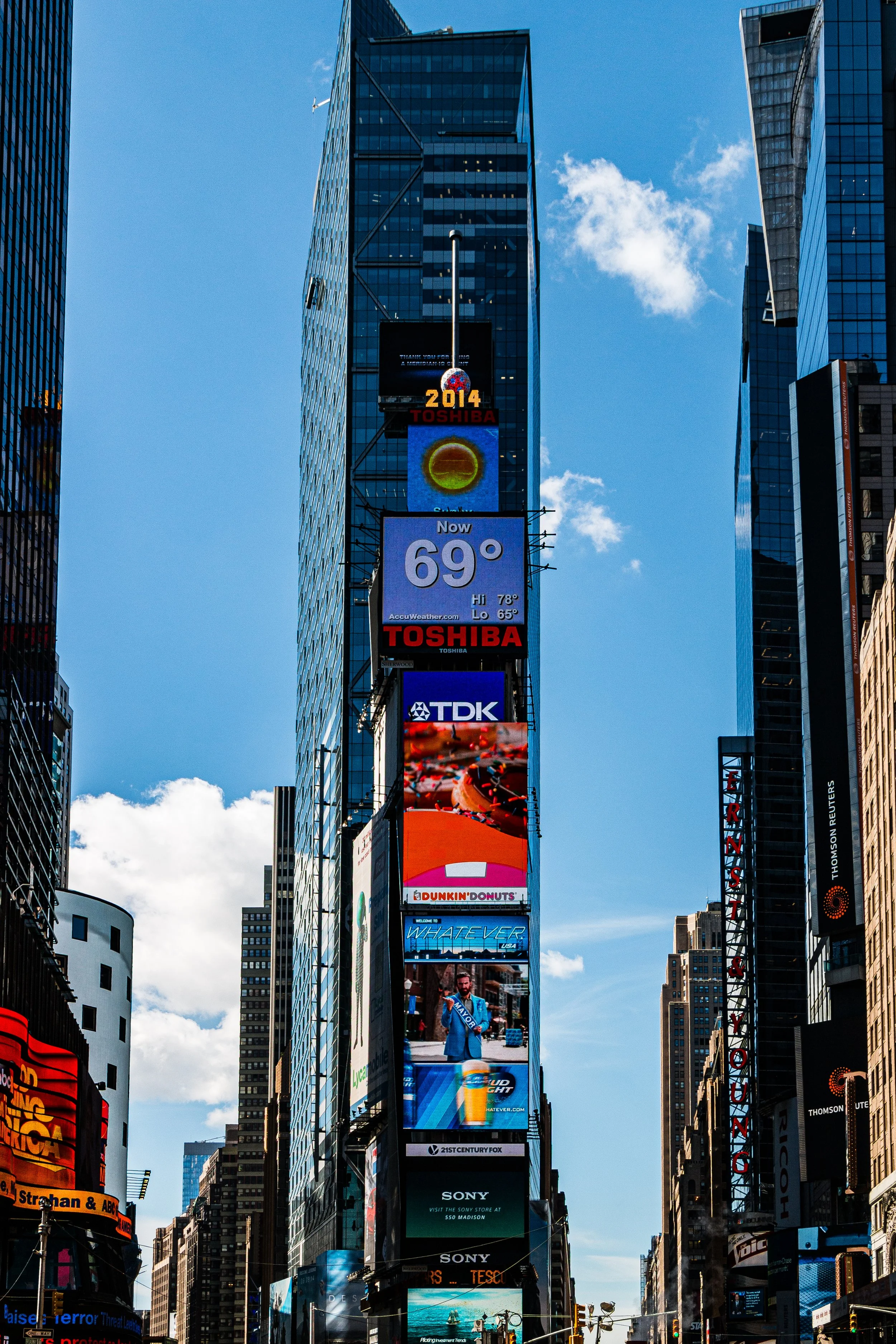 A tall building in Times Square, New York City, displaying various digital billboards including a weather forecast showing 69°F, advertisements for Toshiba, TDK, Dunkin' Donuts, Sony, and other brands, with other skyscrapers and a clear blue sky in the background.