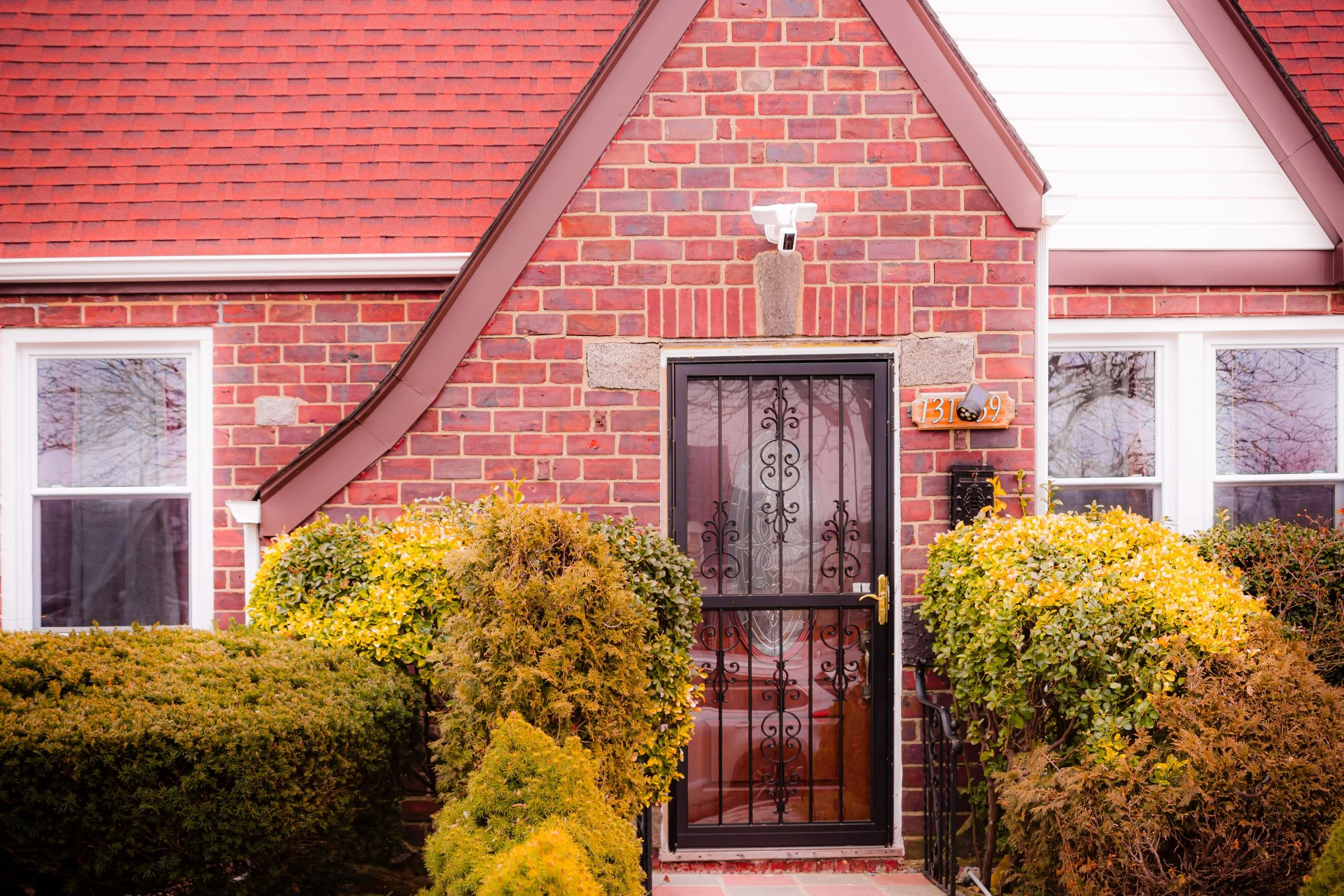 Front door of a brick house covered with bushes and shrubs, with a security camera above.