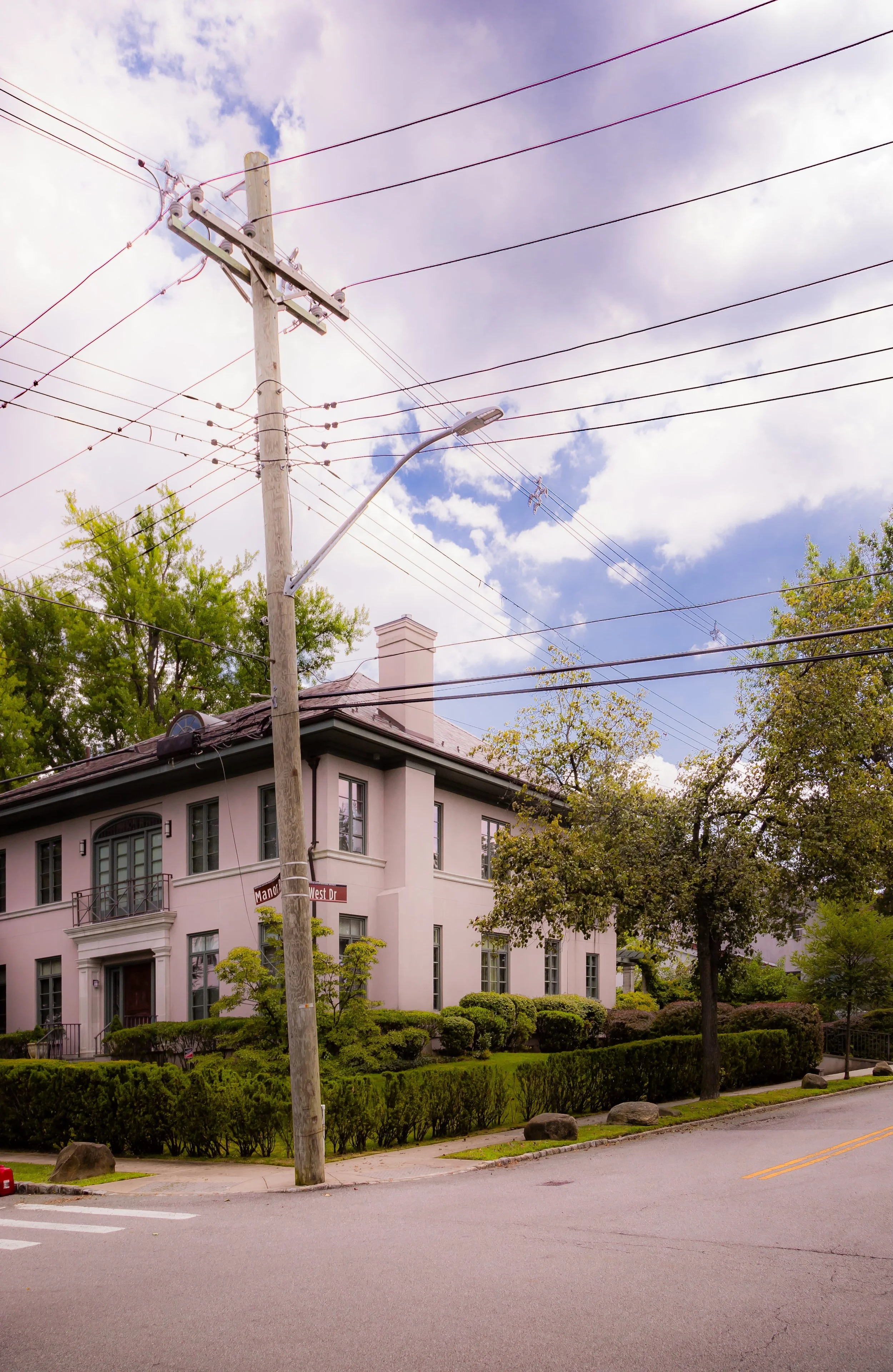 A white two-story house with tall windows, surrounded by green bushes and trees, on a street corner with a wooden utility pole and power lines overhead. The sky is partly cloudy with patches of blue.
