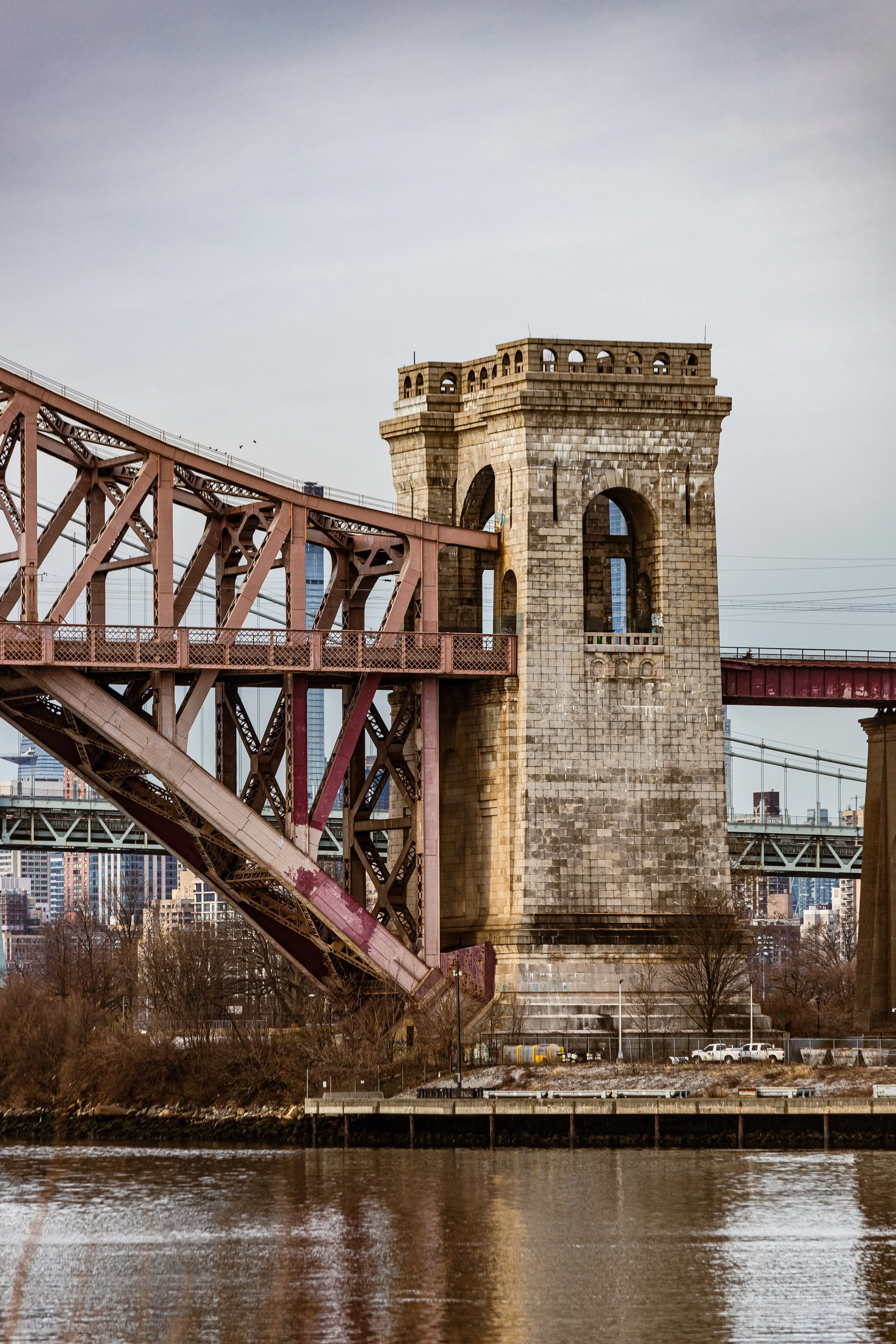 View of a historic stone bridge tower with a large arch, supporting a steel bridge span over a river, with city buildings in the background.