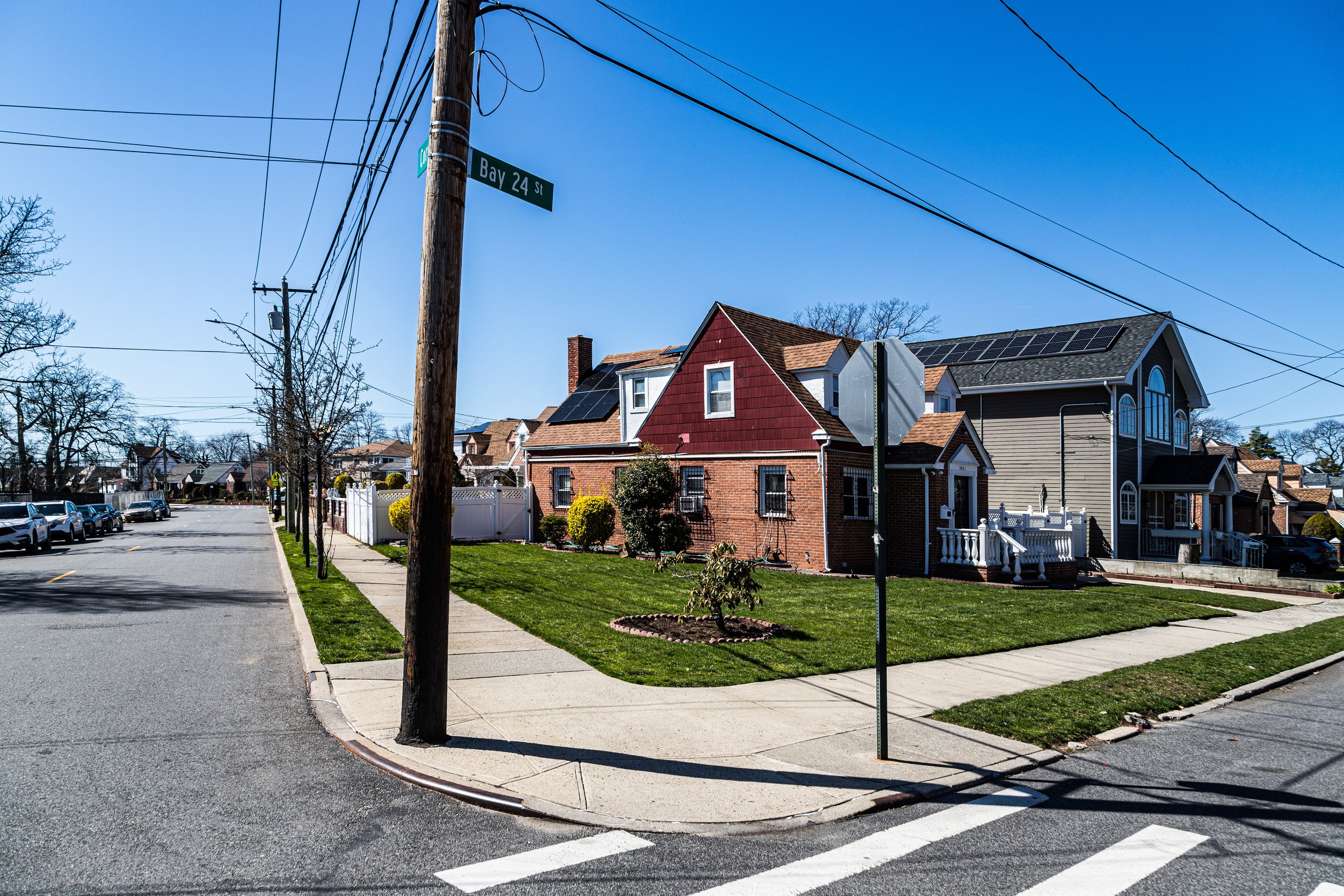 A corner of a residential neighborhood with houses, a sidewalk, a street with parked cars, utility poles, and power lines, under a clear blue sky. The houses have well-maintained lawns and some feature solar panels.