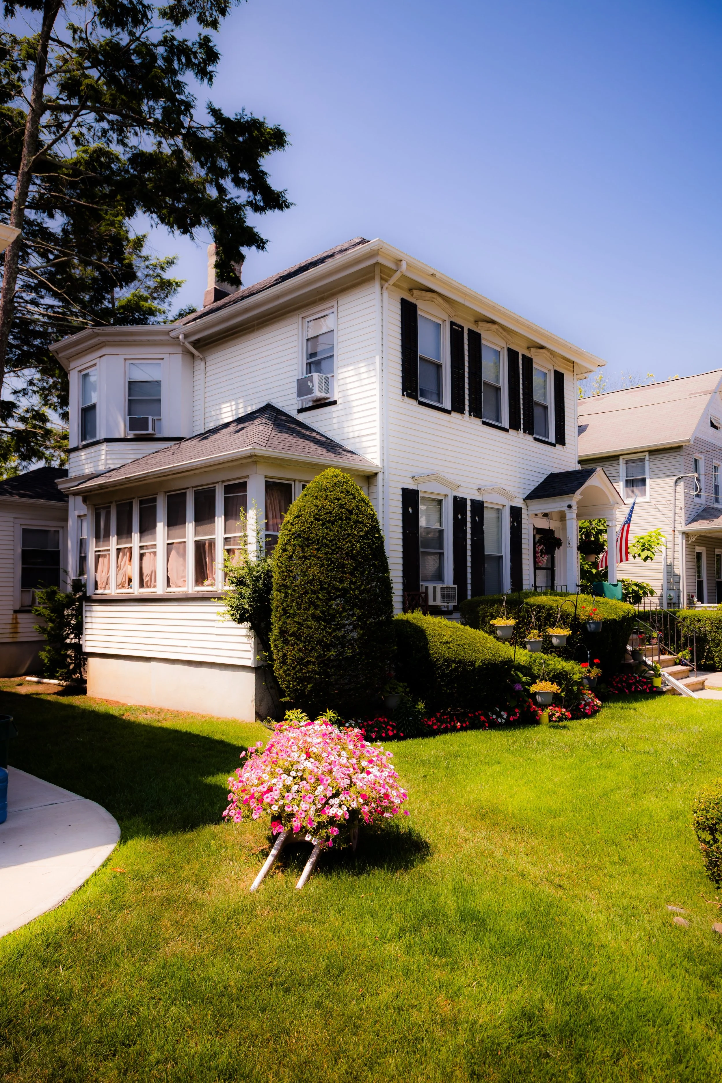 A white house with black shutters and a porch, surrounded by grass, bushes, and flowers, under a clear blue sky.