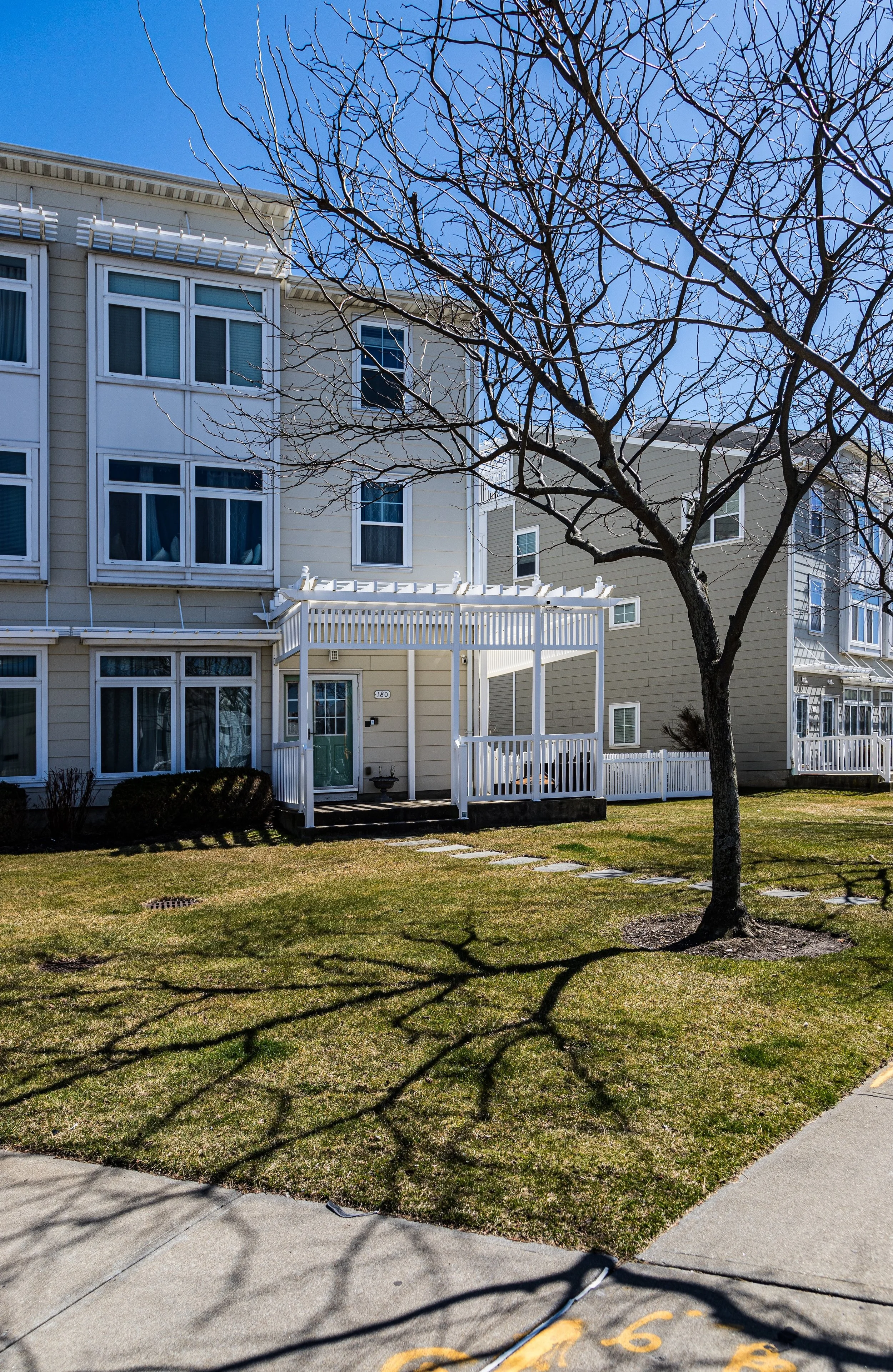 A residential apartment building with a small front porch, surrounded by a well-kept lawn and a leafless tree, under a clear blue sky.